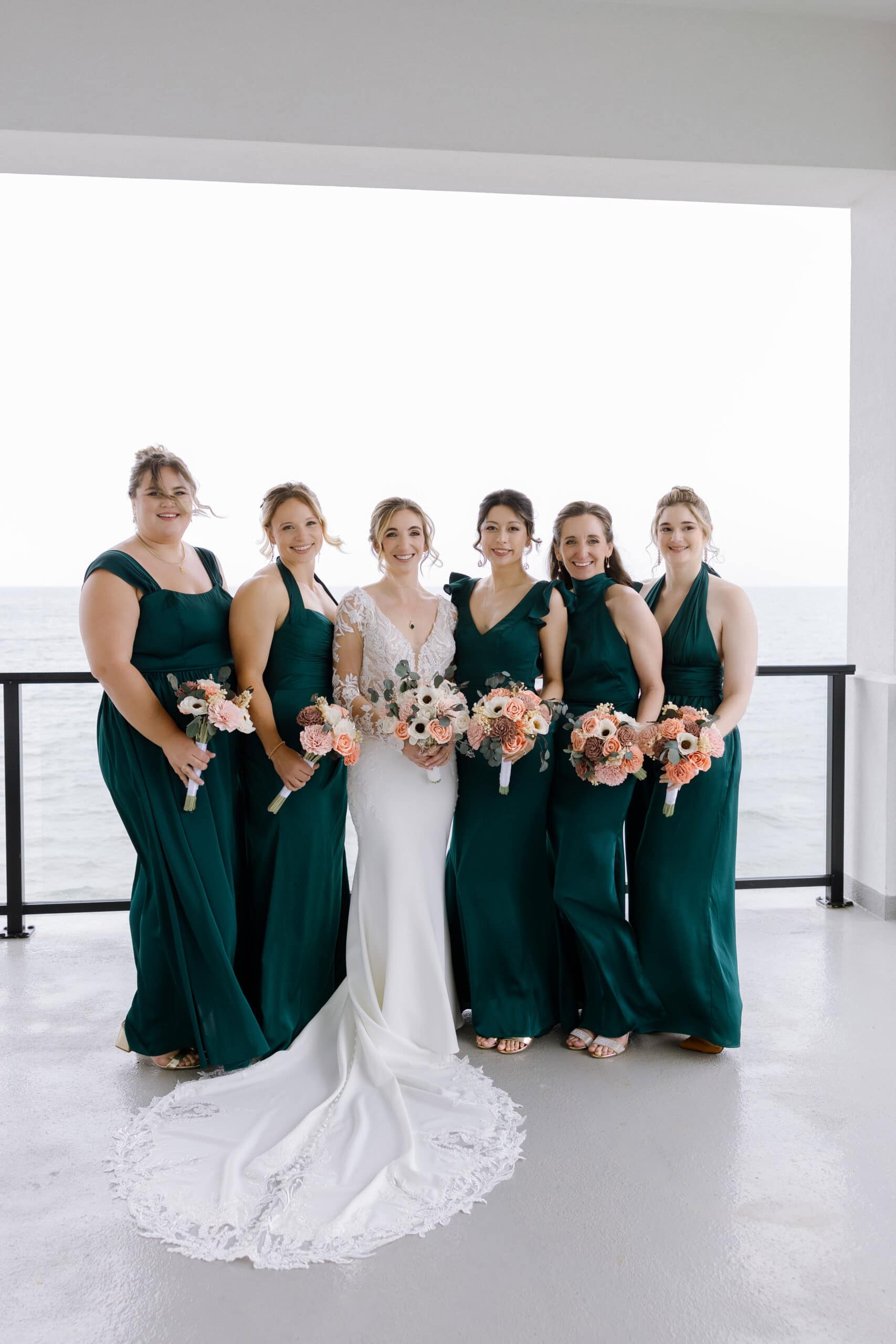 Bride in lace-sleeved white gown with train standing with five bridesmaids in emerald dresses holding bouquets on a balcony overlooking the ocean at Tyde at Walnut Beach, Milford, CT.