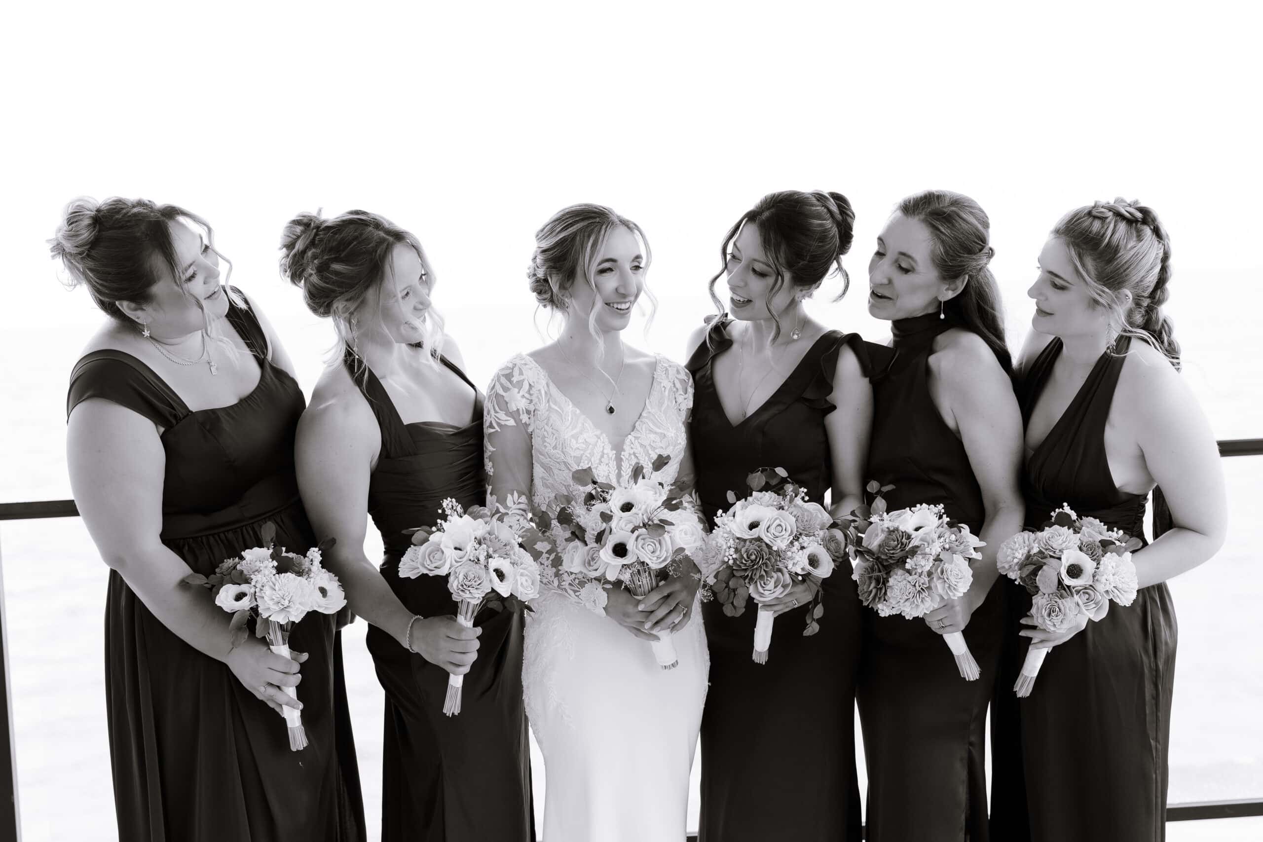 Black and white image of a bride in a lace wedding dress surrounded by five bridesmaids in dark dresses holding bouquets, standing close together at Tyde at Walnut Beach, Milford, CT.