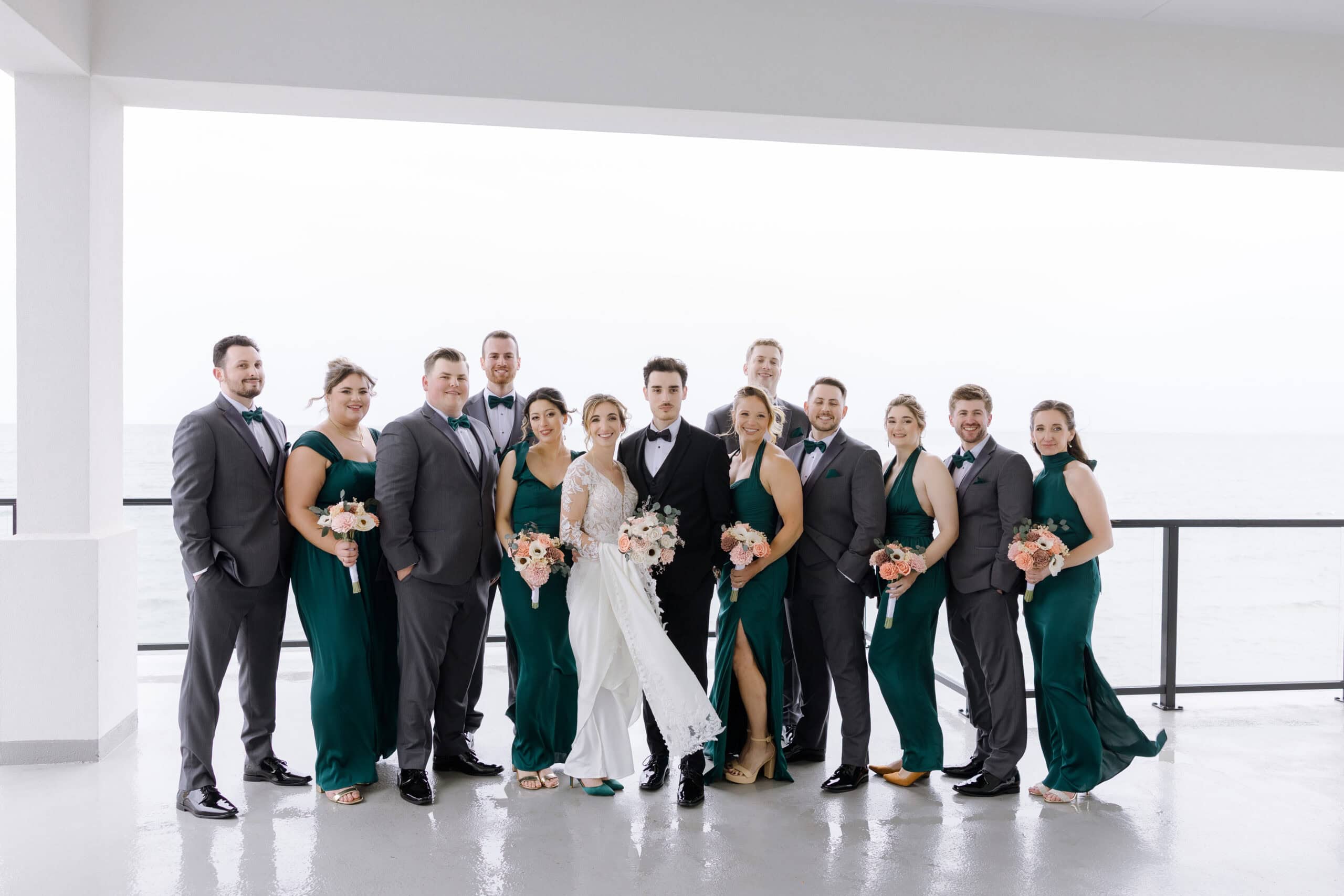 Wedding party in emerald green dresses and gray tuxedos with bow ties standing on a balcony overlooking the ocean at Tyde at Walnut Beach, Milford, CT.
