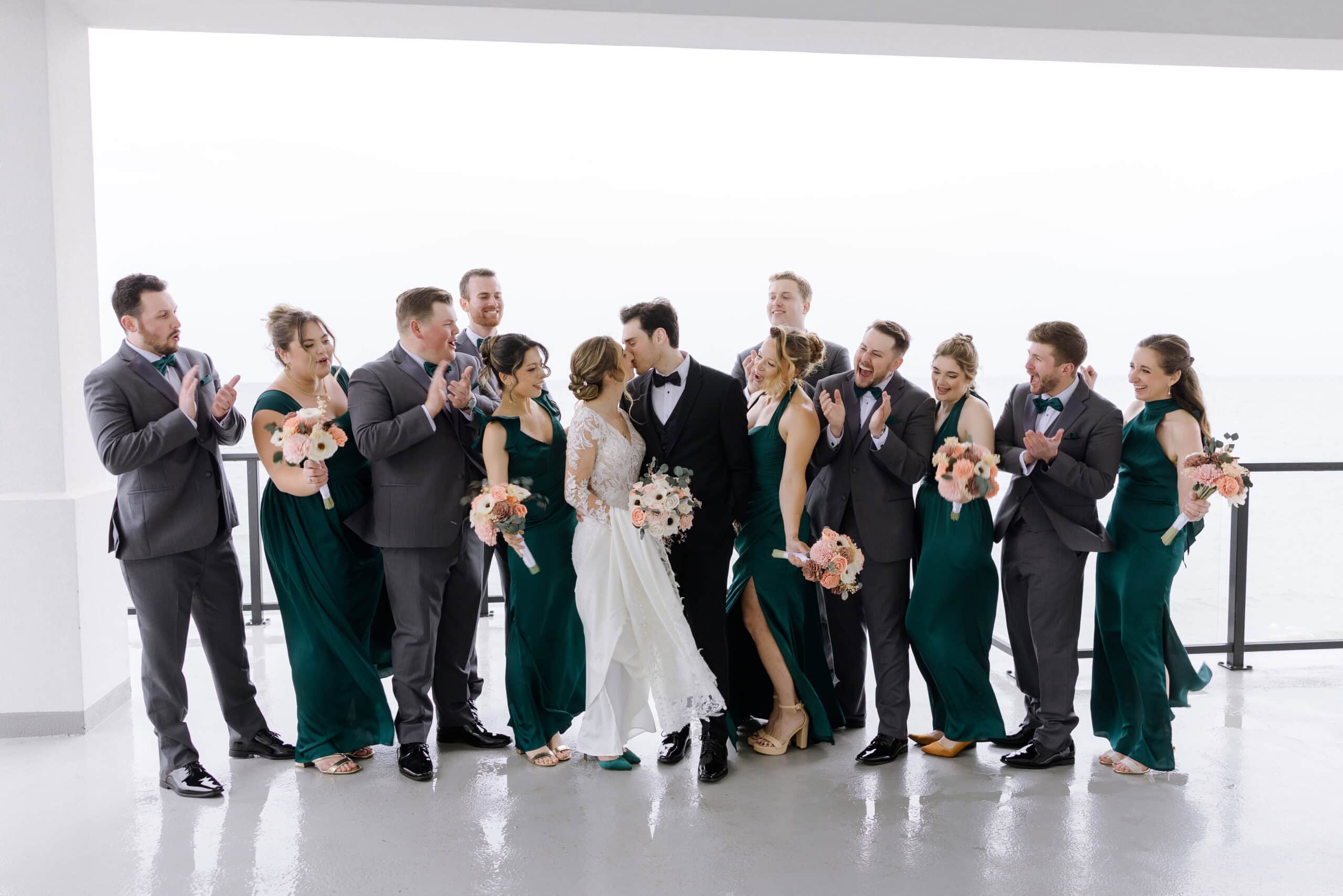 Wedding party in emerald green dresses and charcoal suits applauding as bride and groom kiss on balcony overlooking the sea at Tyde at Walnut Beach, Milford, CT.