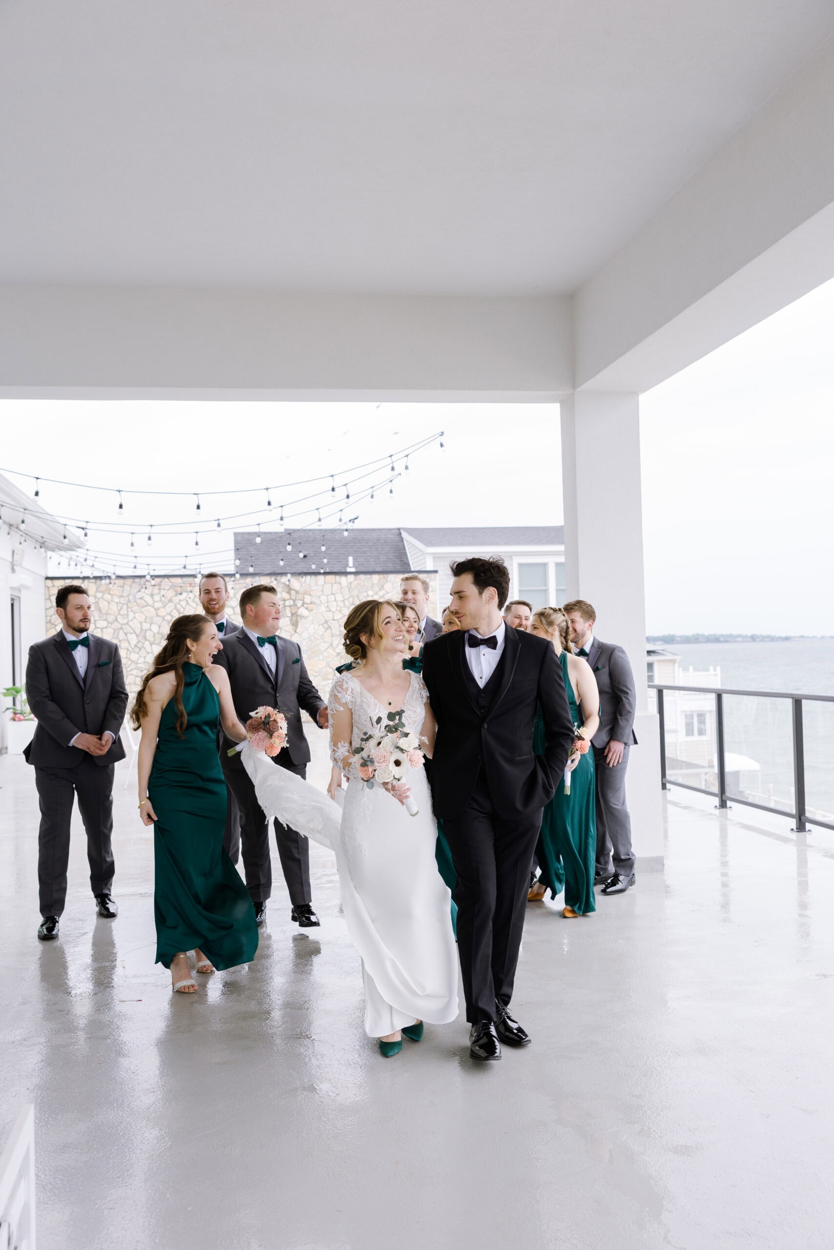 Bride in lace gown and groom in black tuxedo walking with bridesmaids in emerald green dresses and groomsmen in gray suits on terrace overlooking water at Tyde at Walnut Beach, Milford, CT.
