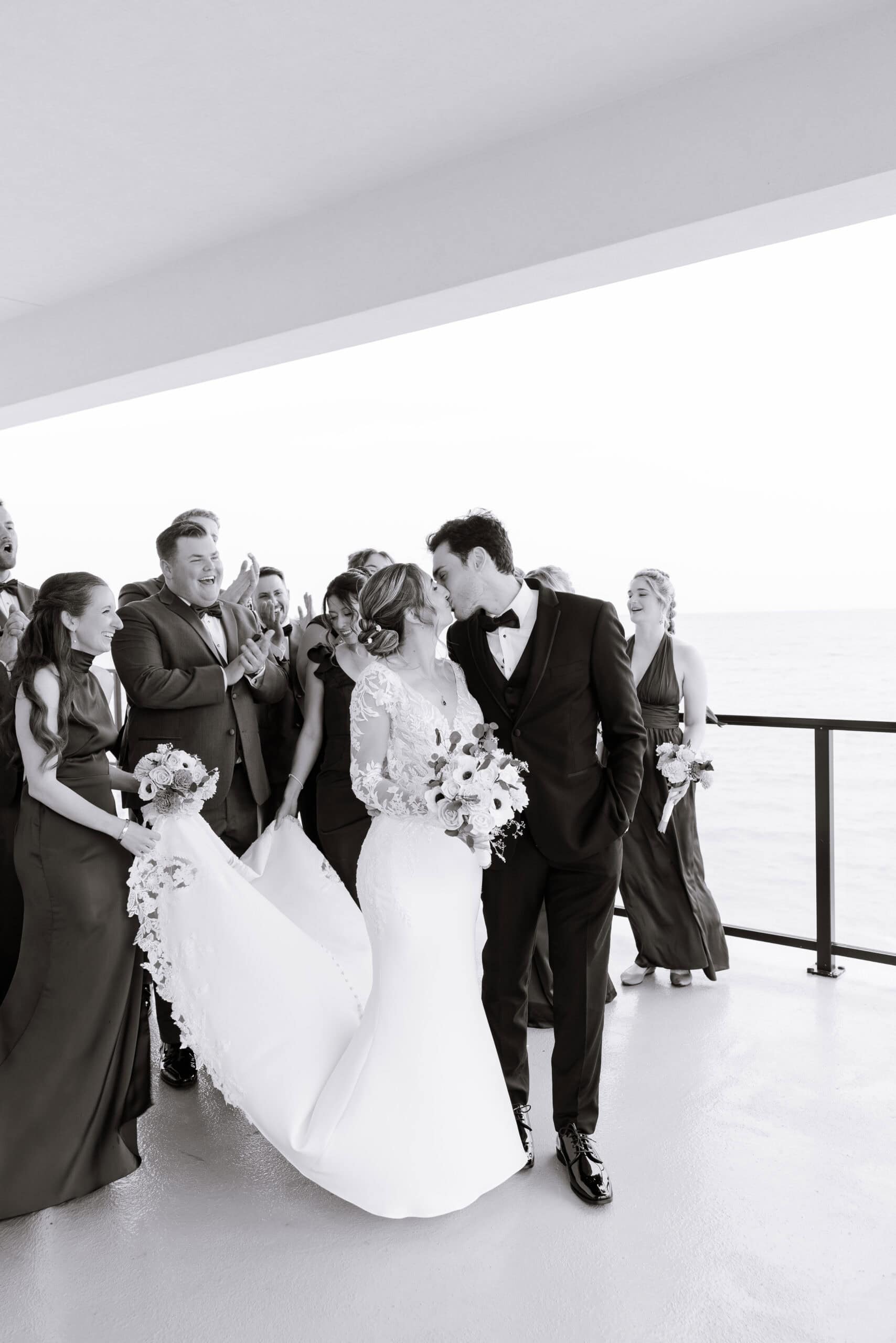 Bride and groom kissing on balcony with bridal party applauding, overlooking ocean at Tyde at Walnut Beach, Milford, CT.