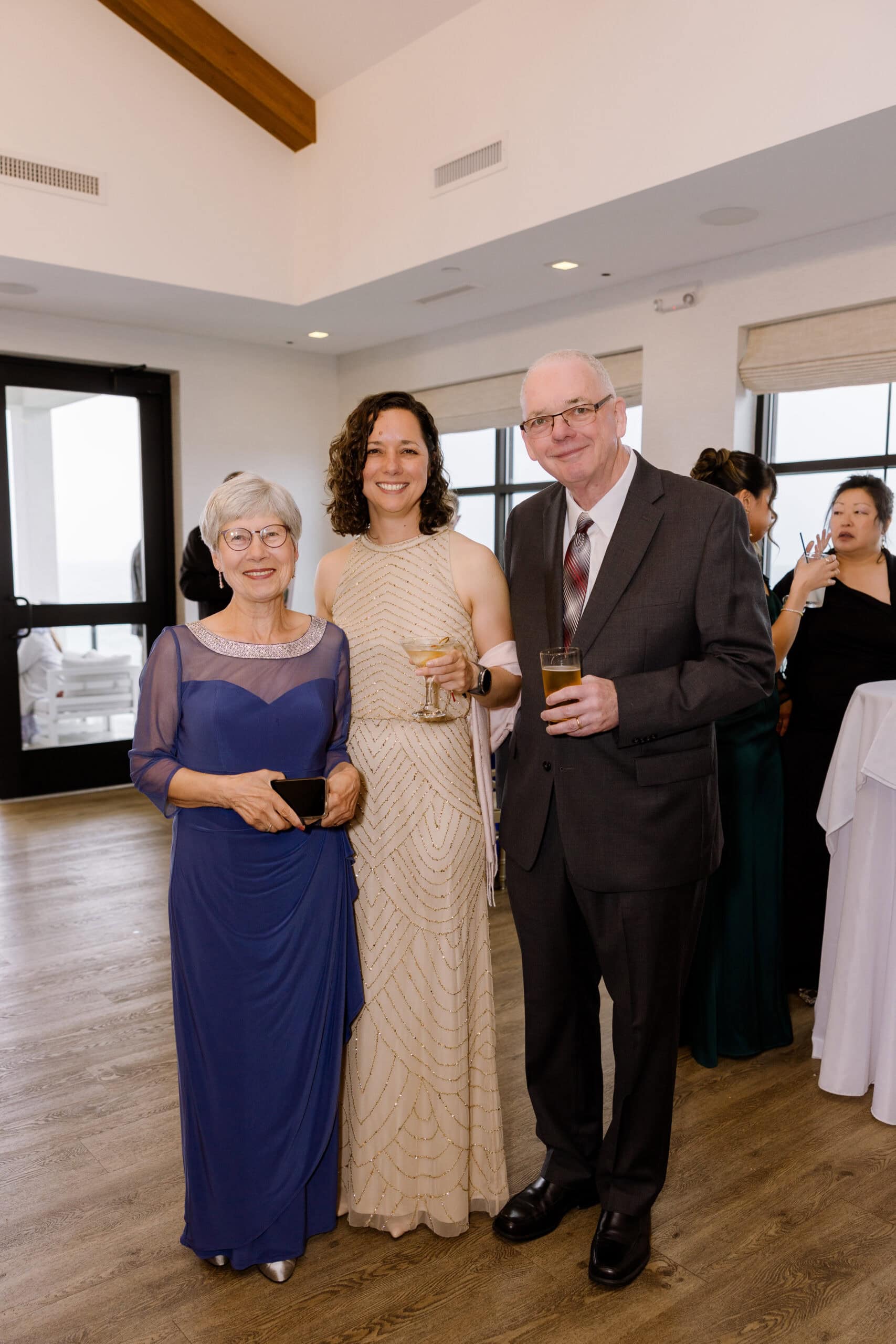Three elegantly dressed guests at a wedding reception inside Tyde at Walnut Beach, Milford, CT, holding drinks and smiling.