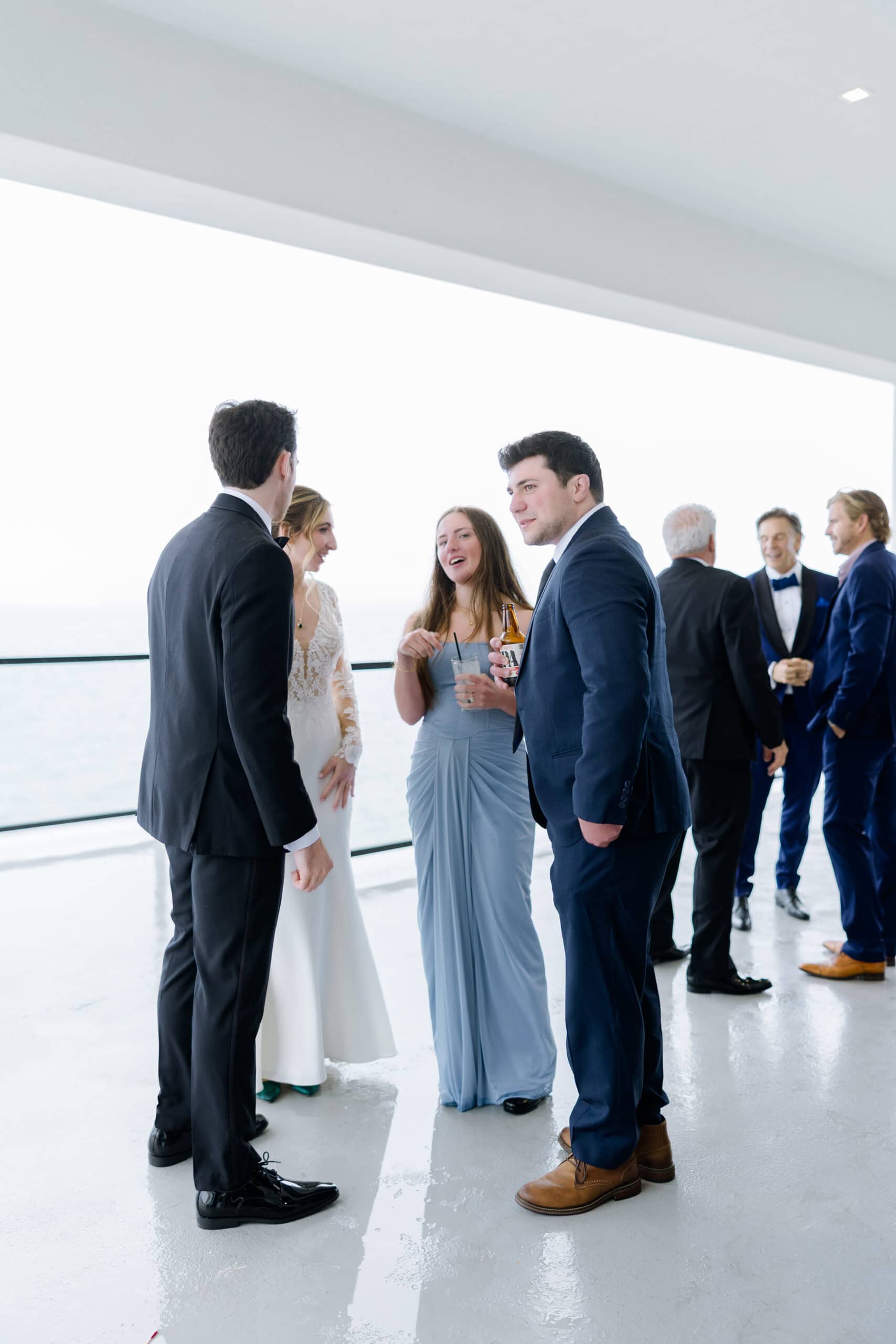 Wedding guests in formal attire conversing on a bright terrace overlooking the ocean at Tyde at Walnut Beach, Milford, CT.