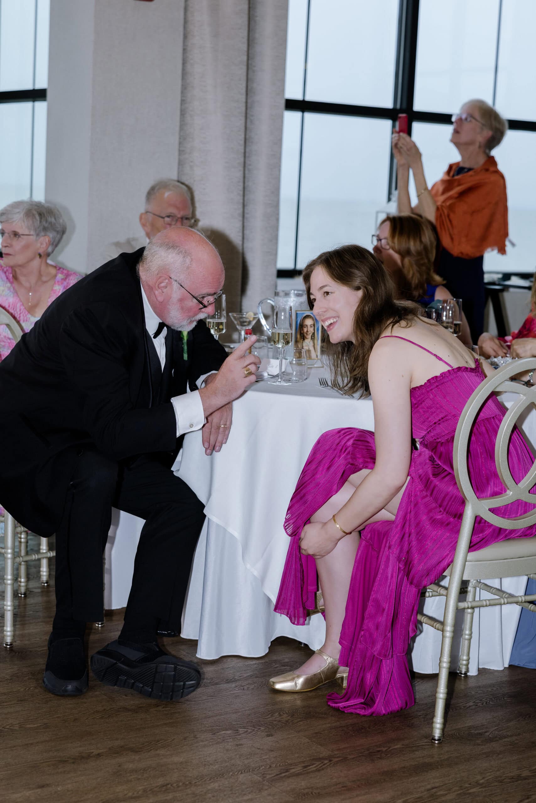 Elderly man in tuxedo leaning forward to speak with young woman in magenta dress seated at a wedding reception table with glassware and framed photo, indoor venue with large windows.