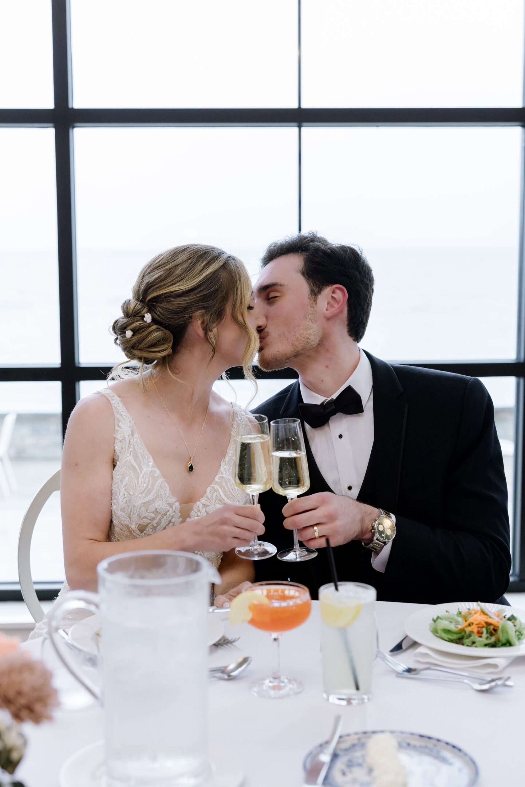 Bride and groom in elegant attire kissing while holding champagne glasses near a large window overlooking water at Tyde at Walnut Beach, Milford, CT.