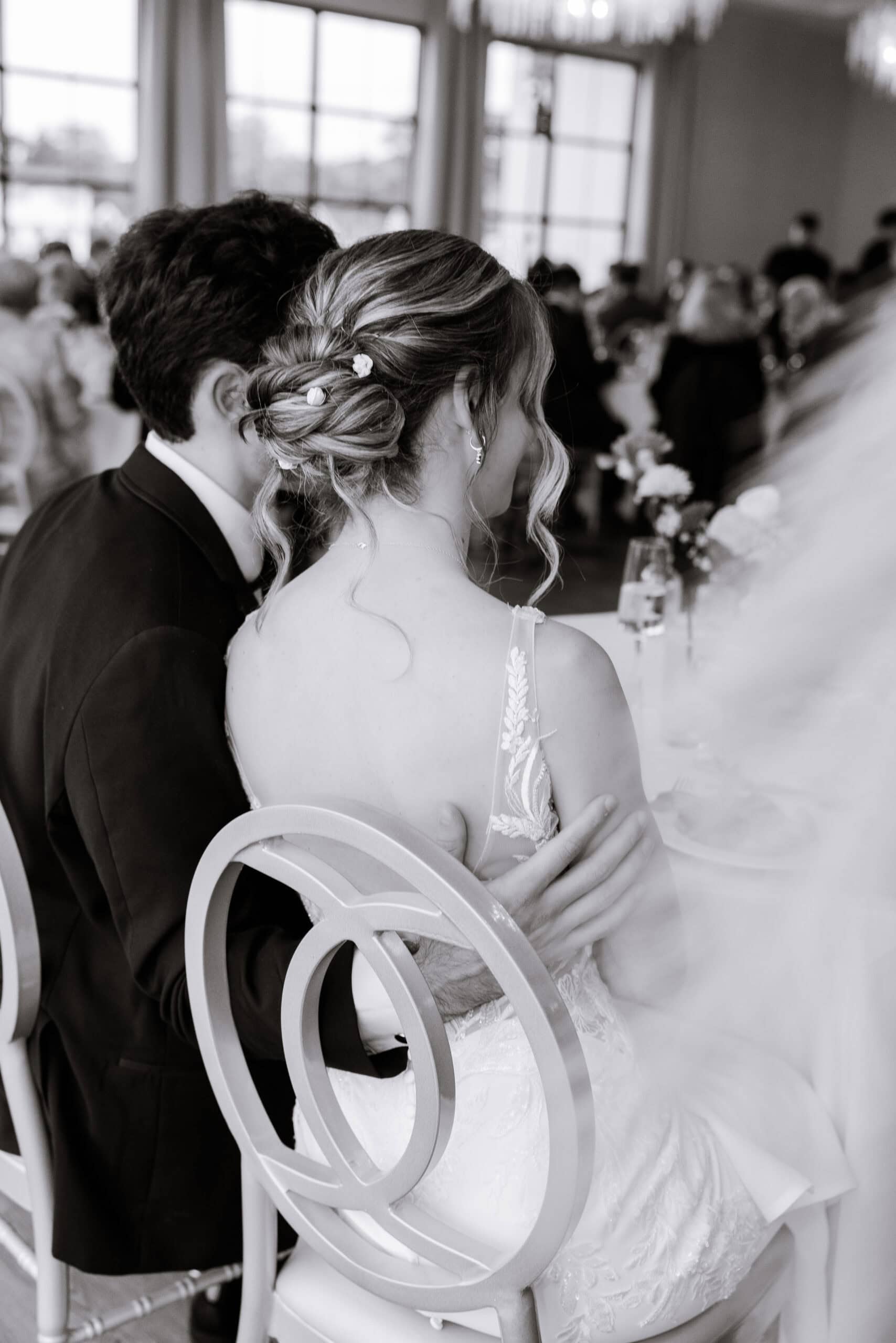 Black and white photo of a couple seated closely at a wedding reception, the groom's hand resting on the bride's back, highlighting her lace dress and floral hair accessories.