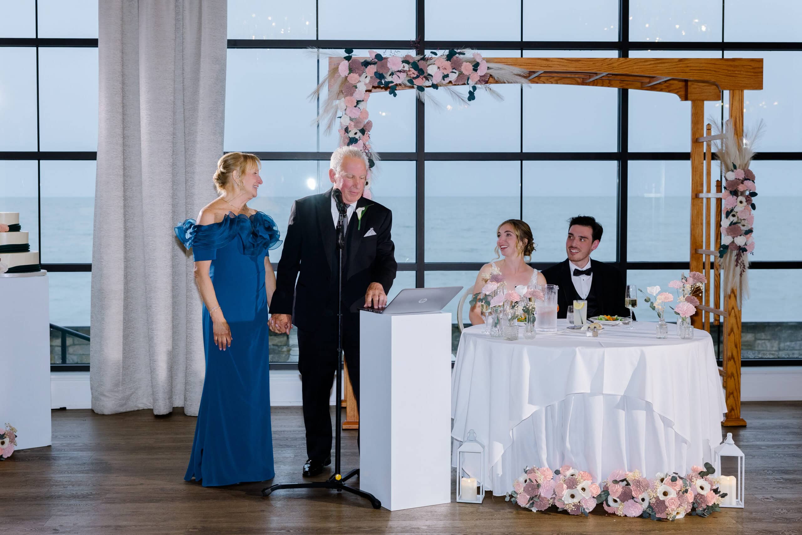 Couple seated at a white-clothed table adorned with pink floral arrangements, listening to a man in a tuxedo giving a speech beside a woman in a blue gown, with large windows showing ocean view in background.