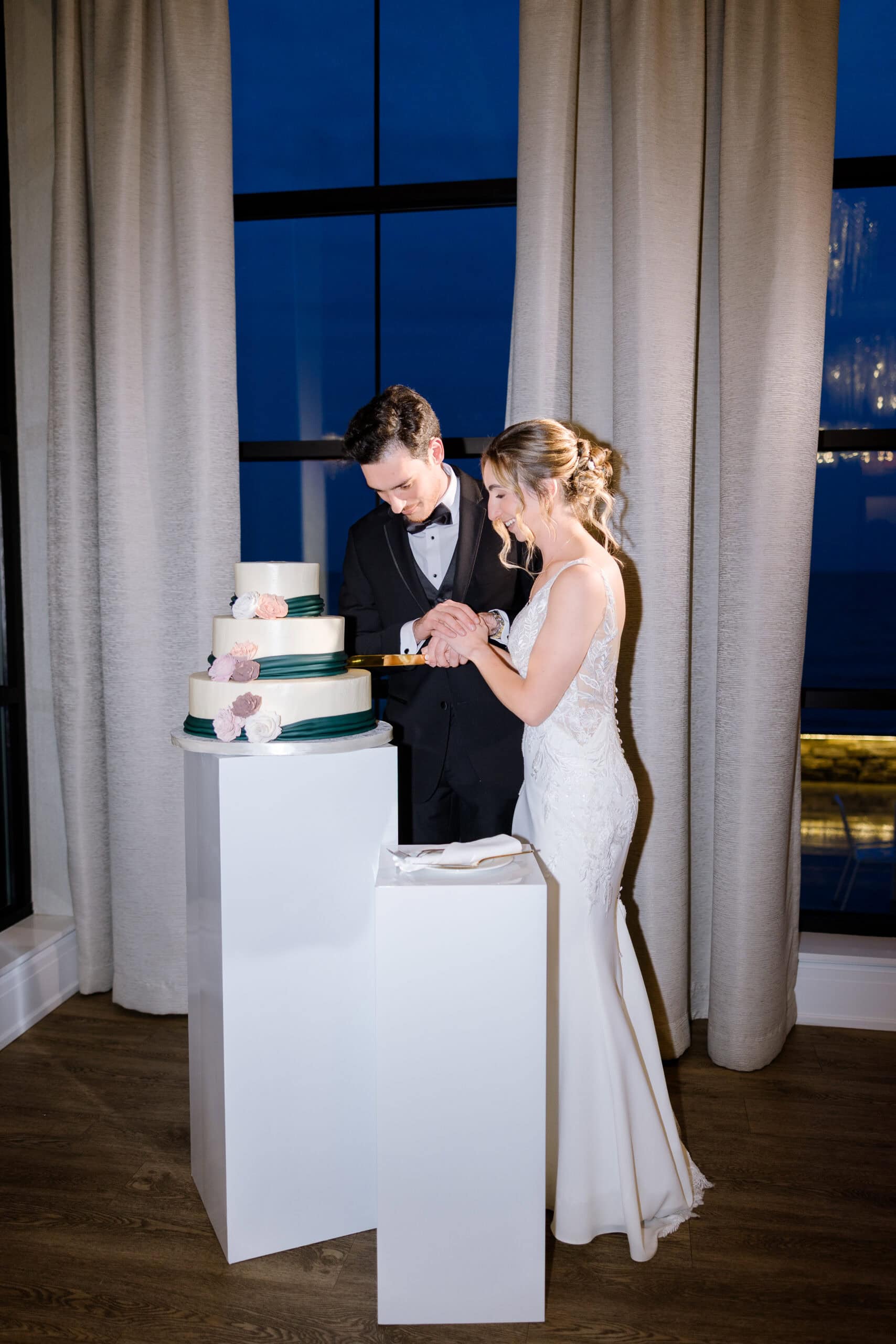 Bride and groom cutting a three-tiered wedding cake decorated with green ribbons and flowers, standing by large windows with beige curtains at night.