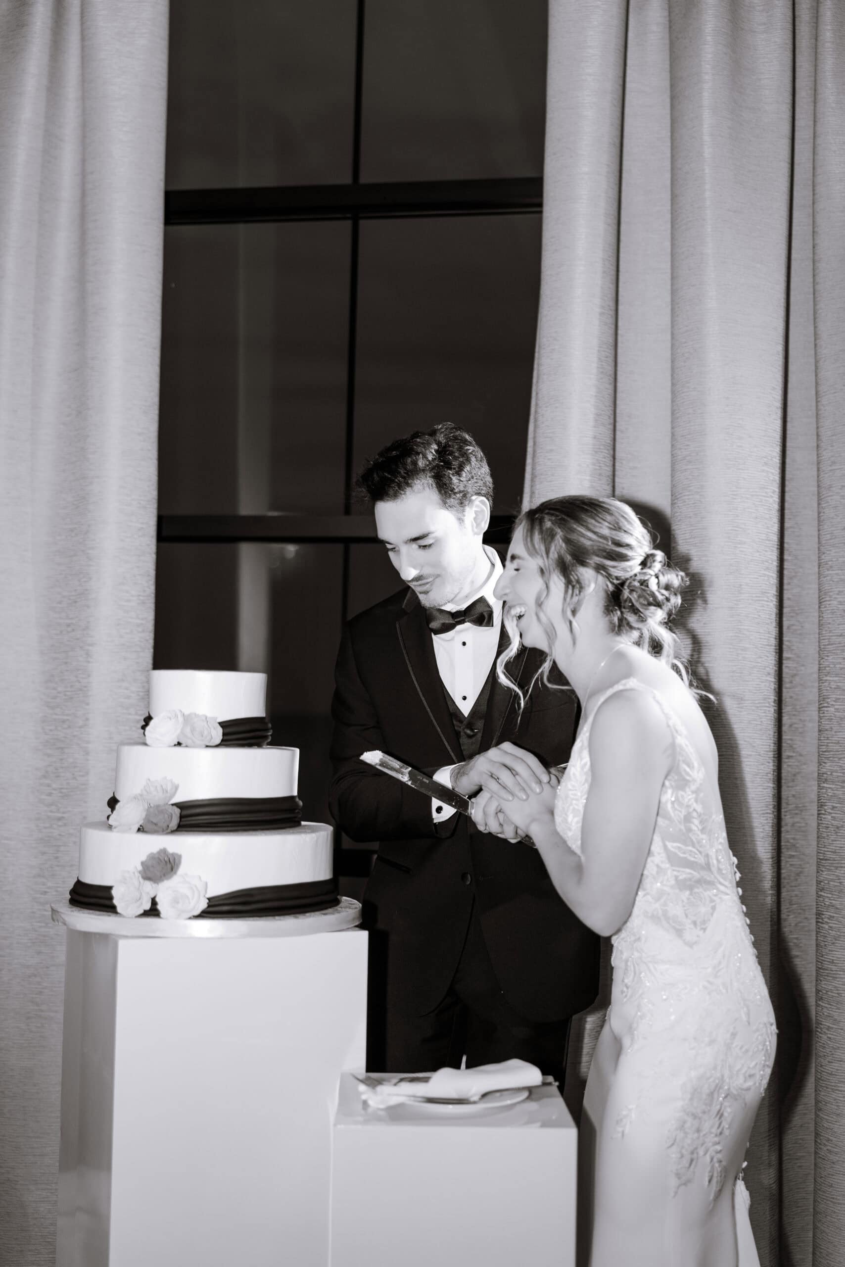 Bride and groom cutting a three-tier wedding cake adorned with white flowers and black ribbon, standing by tall curtains at Tyde at Walnut Beach, Milford, CT.