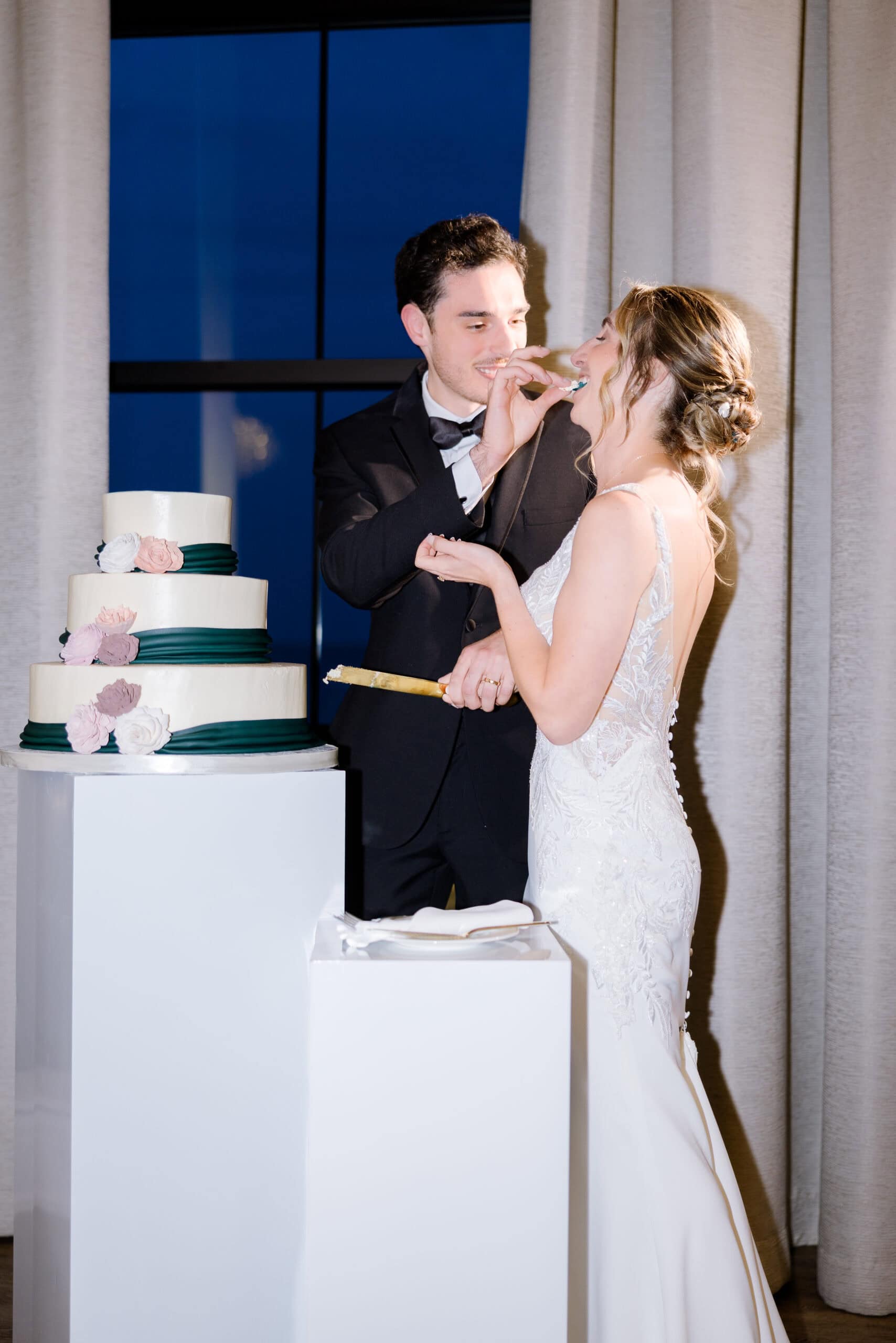 Groom in black tuxedo feeding bride in lace wedding gown a piece of wedding cake beside a three-tiered cake with floral decorations at Tyde at Walnut Beach.