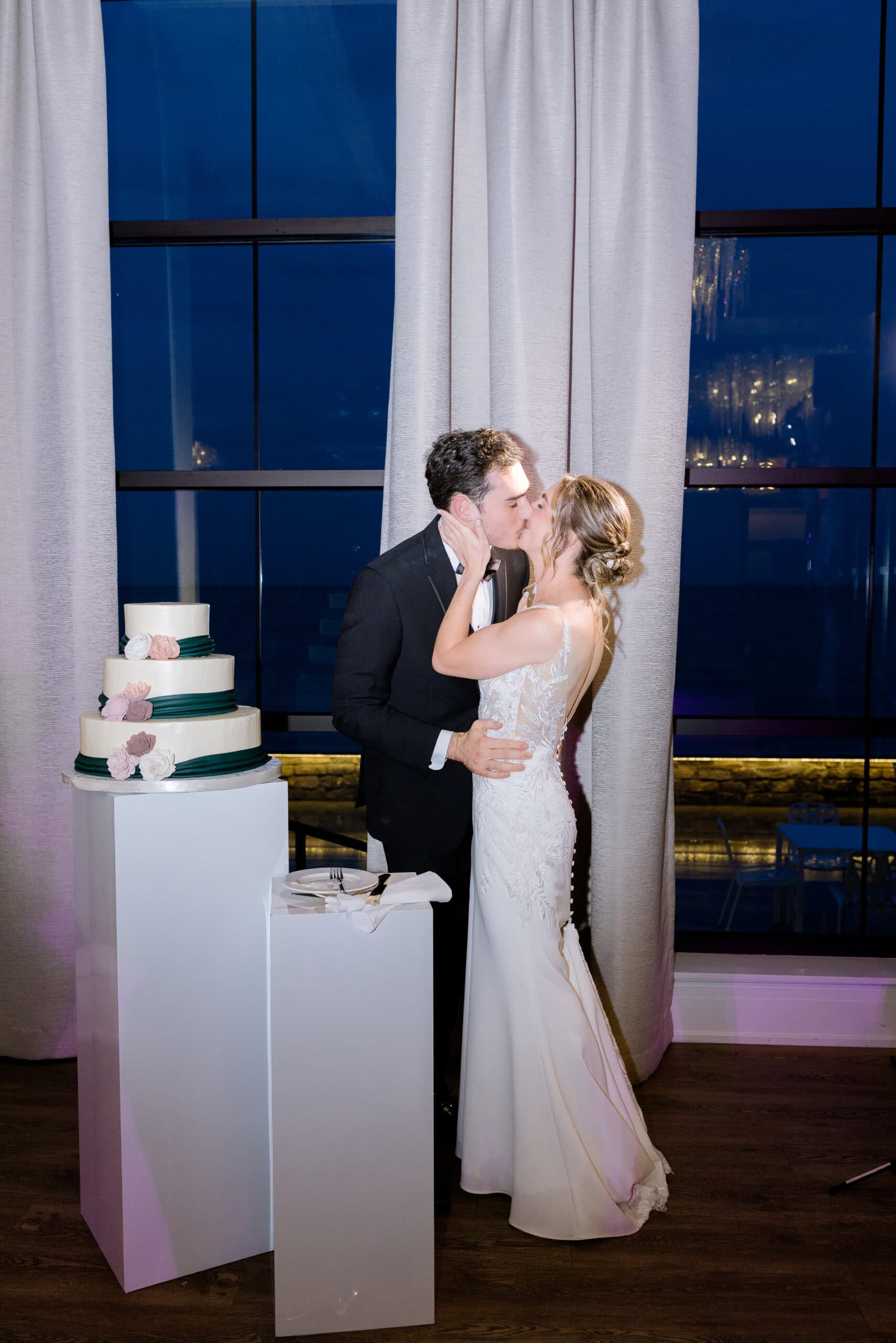 Bride and groom kissing next to a three-tiered wedding cake with floral decorations, in front of large windows with curtains at Tyde at Walnut Beach, Milford, CT.