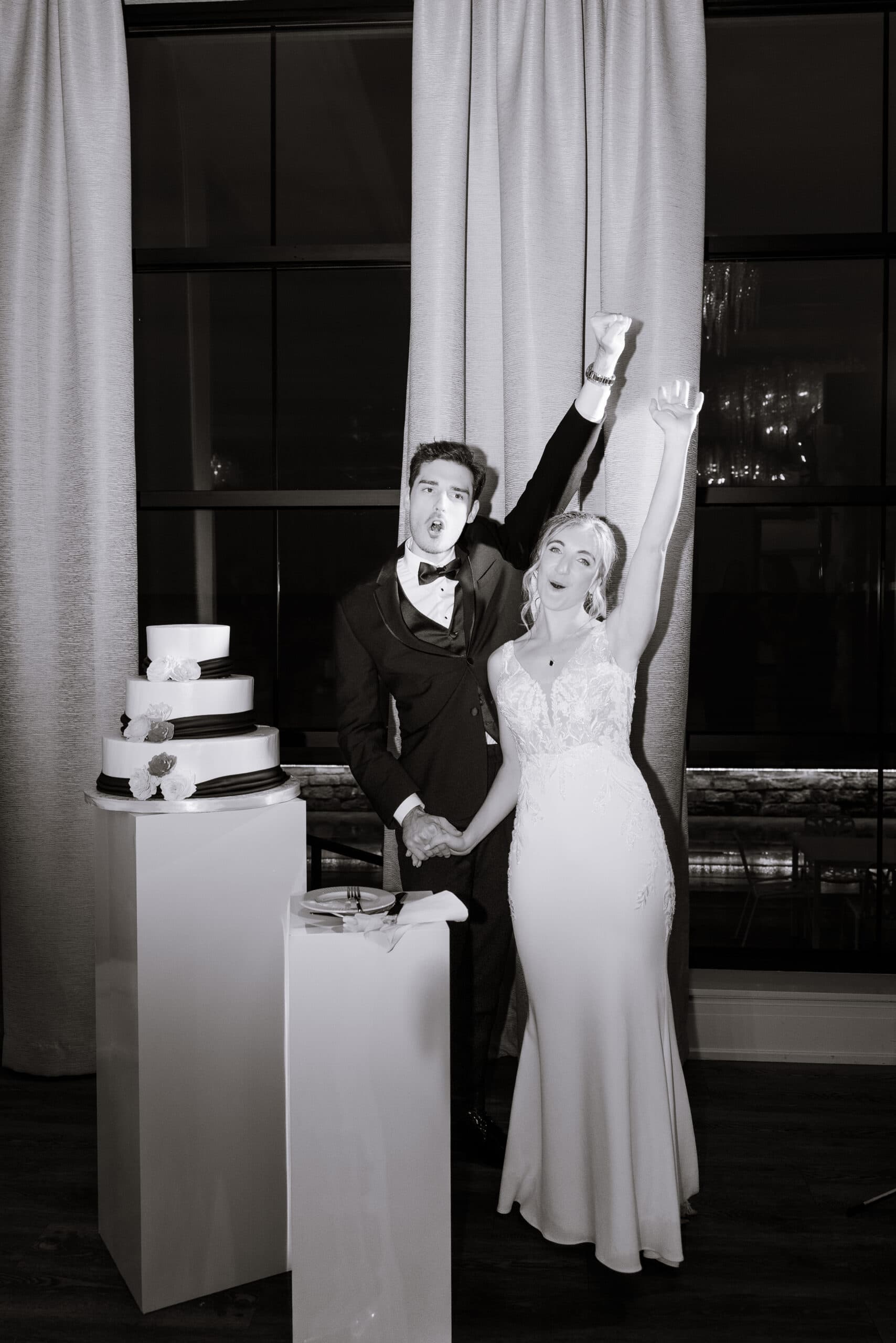 Bride and groom in elegant attire raising arms in celebration next to a four-tier wedding cake on white pedestals, indoor setting with large window and curtains.