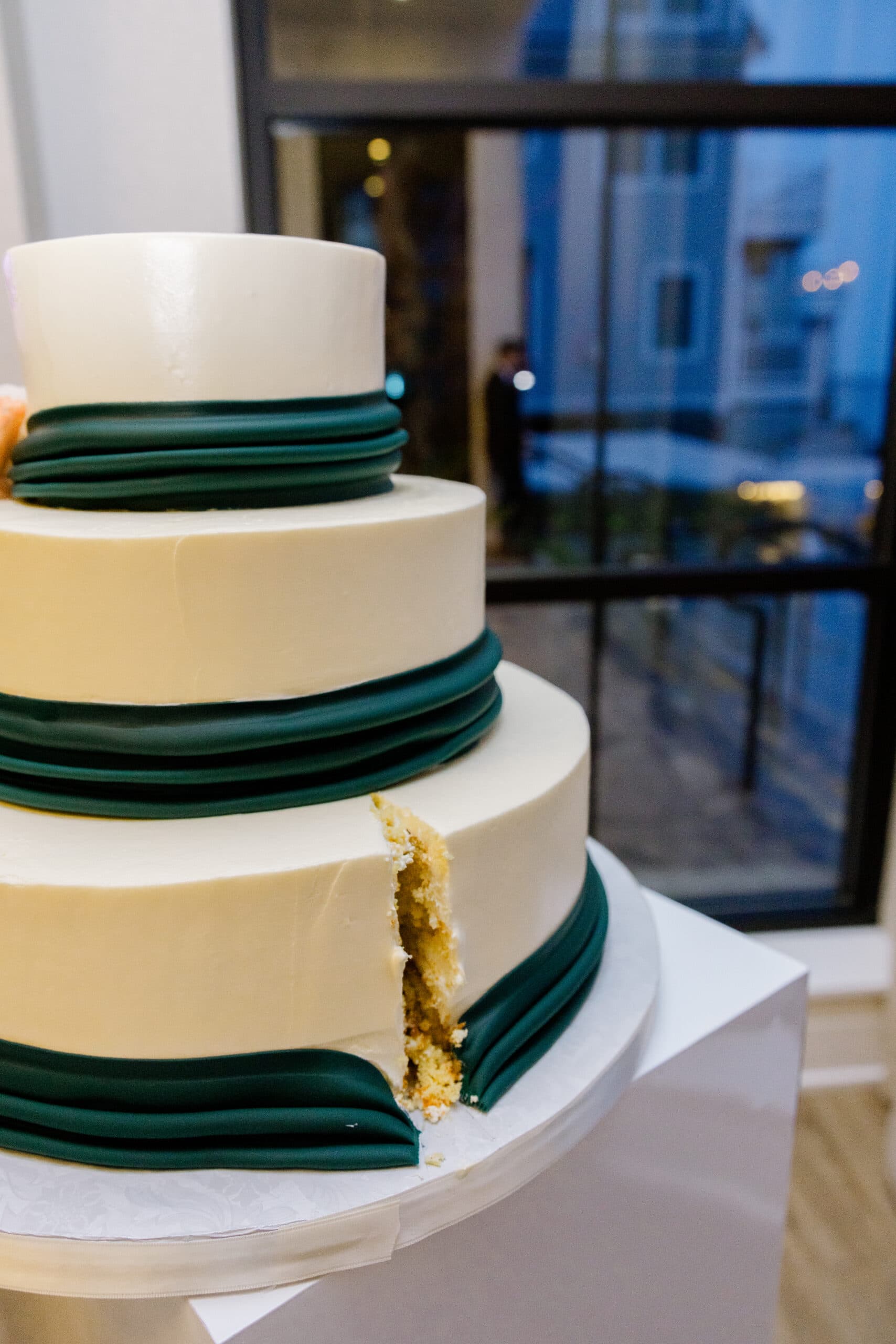 Close-up of a three-tier white wedding cake with dark green fondant bands and a slice cut out, set against a windowed background at Tyde at Walnut Beach, Milford, CT.