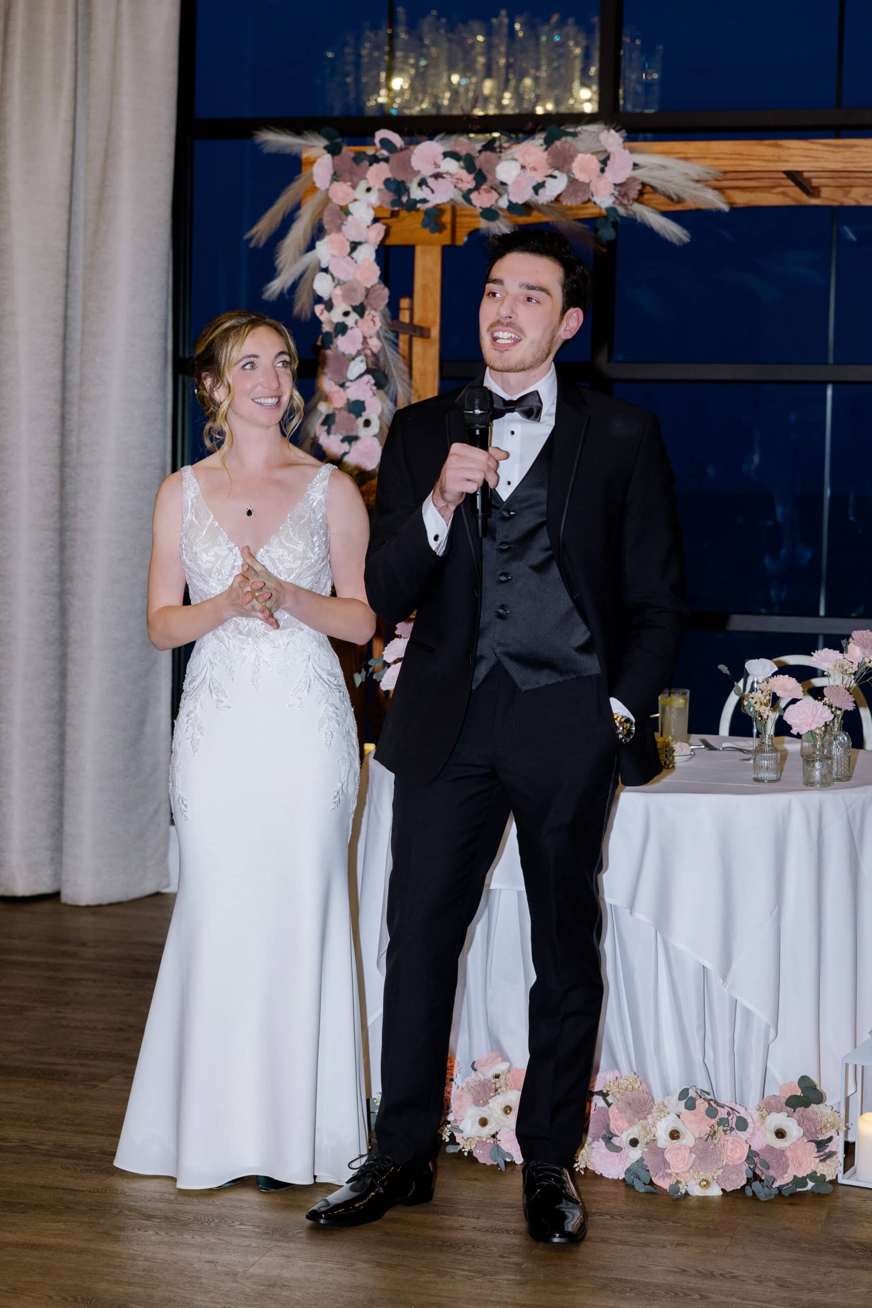 Bride in lace wedding dress and groom in black tuxedo giving a speech beside a floral arch at Tyde at Walnut Beach venue.