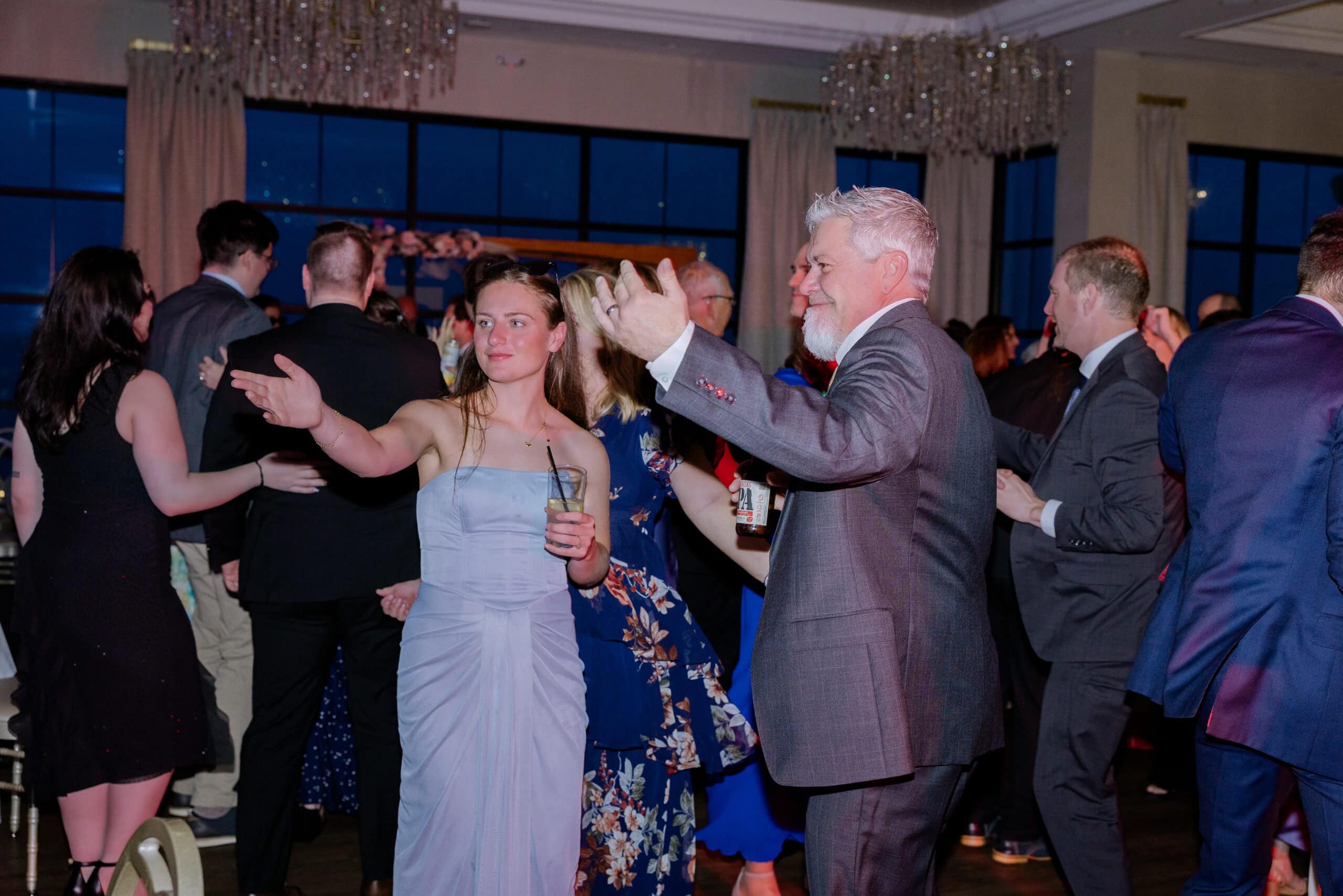 Guests dancing indoors at Tyde at Walnut Beach venue in Milford, CT, with a woman in a pale lilac dress holding a drink and a man in a gray suit holding a beer.