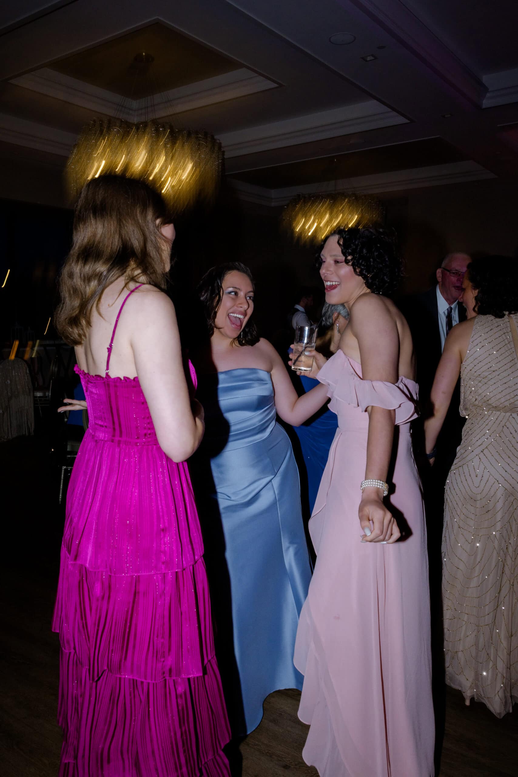 Three women in pink, blue, and magenta gowns laughing and dancing indoors under glowing chandeliers at Tyde at Walnut Beach, Milford, CT.