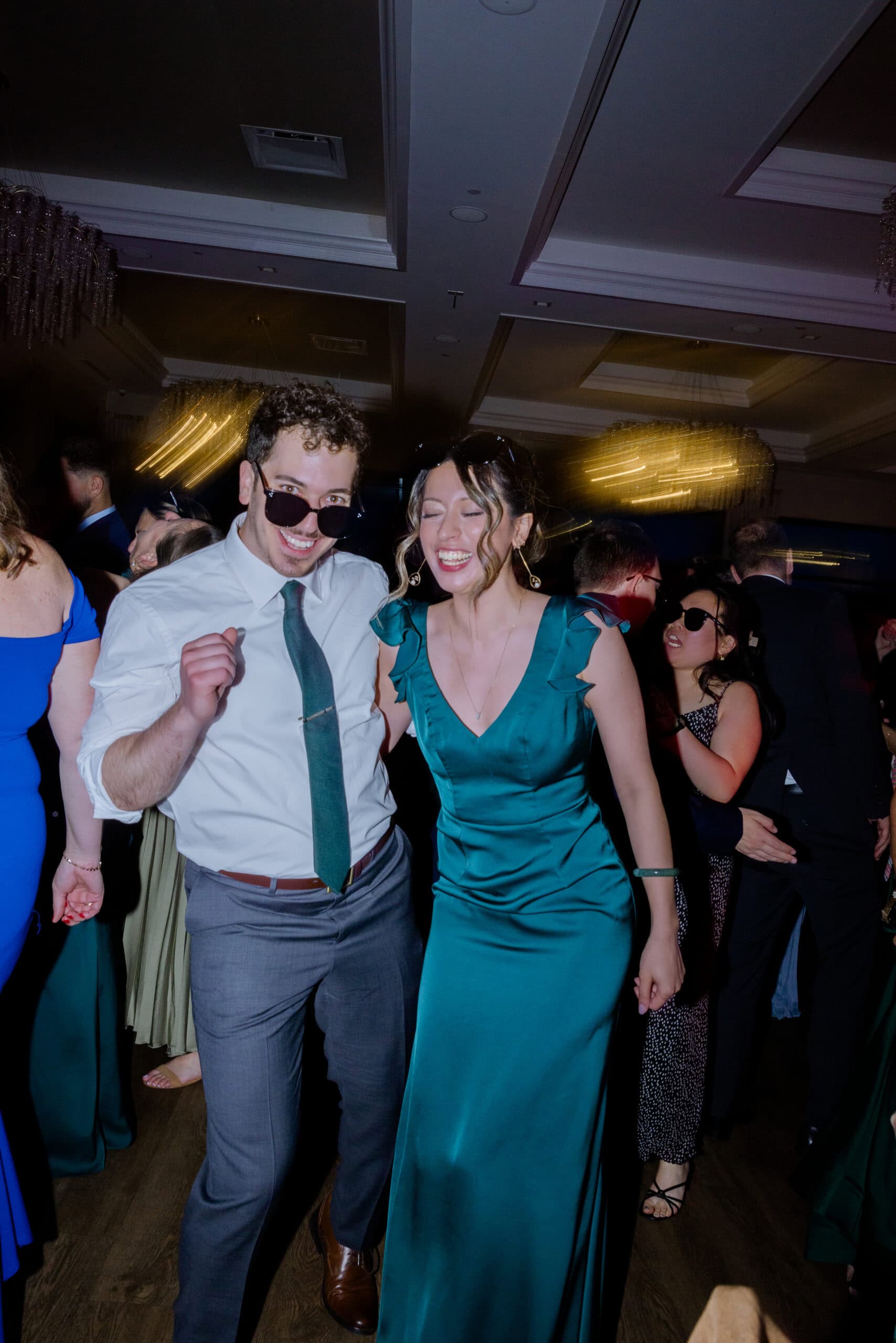 Man and woman dancing indoors at a wedding reception, both wearing sunglasses; woman in emerald green satin dress, man in white shirt with green tie.