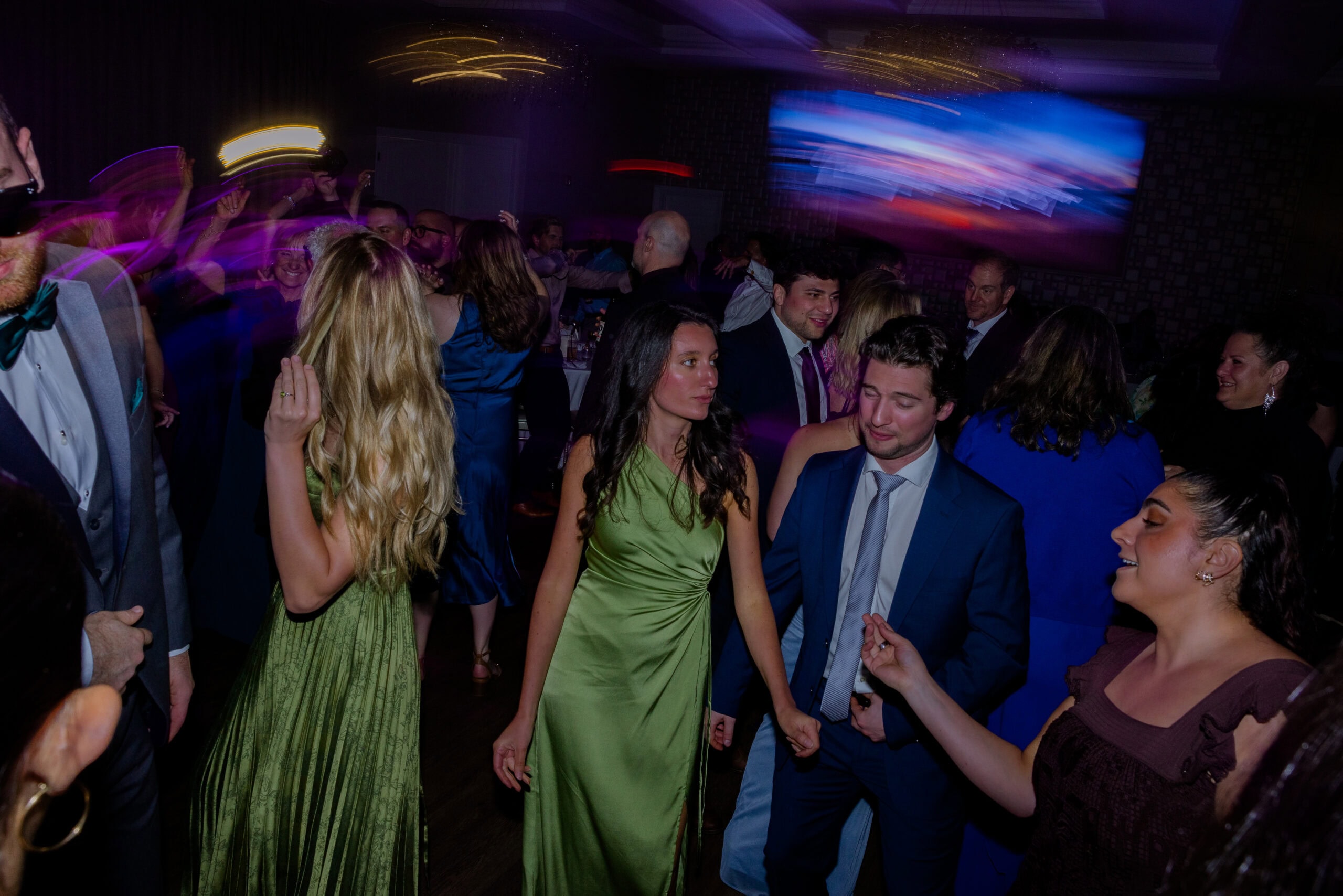 Crowd of elegantly dressed guests dancing and socializing in a dimly lit room with colorful motion blur effects at Tyde at Walnut Beach, Milford, CT.