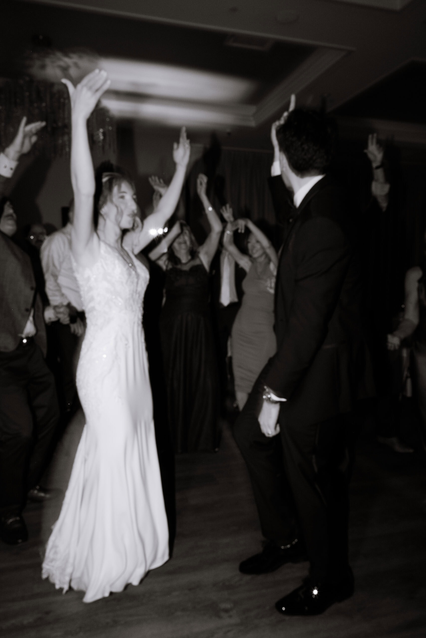 Black and white photo of bride and groom dancing with raised arms surrounded by guests at a wedding reception.