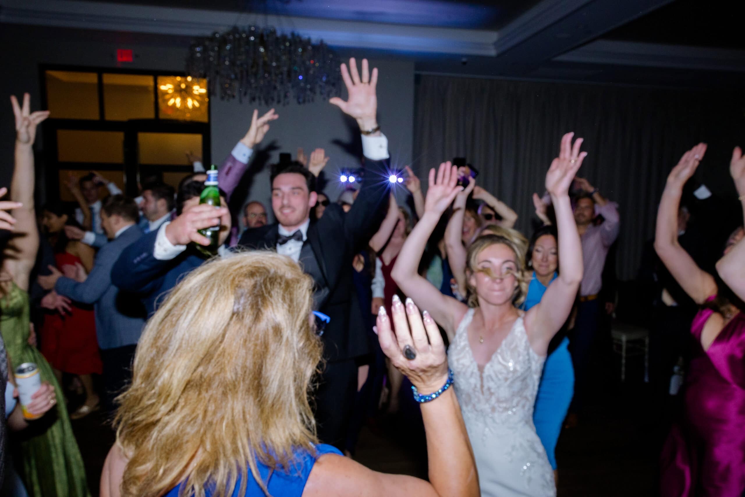 Wedding guests dancing energetically with raised hands in a dimly lit reception hall at Tyde at Walnut Beach, Milford, CT.