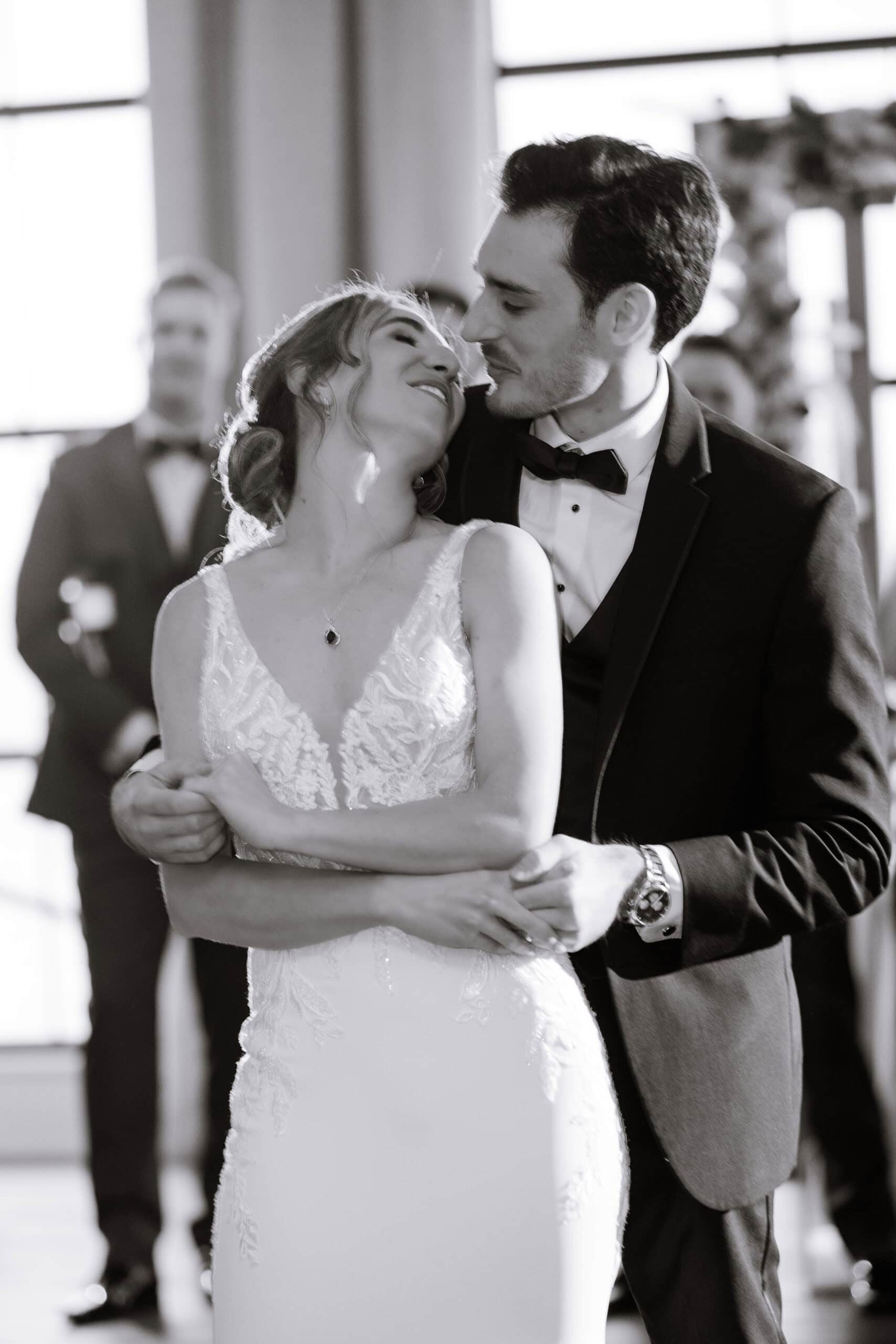 Black and white photo of a bride and groom in an intimate embrace during their wedding, with the groom holding the bride from behind and both gazing affectionately at each other.
