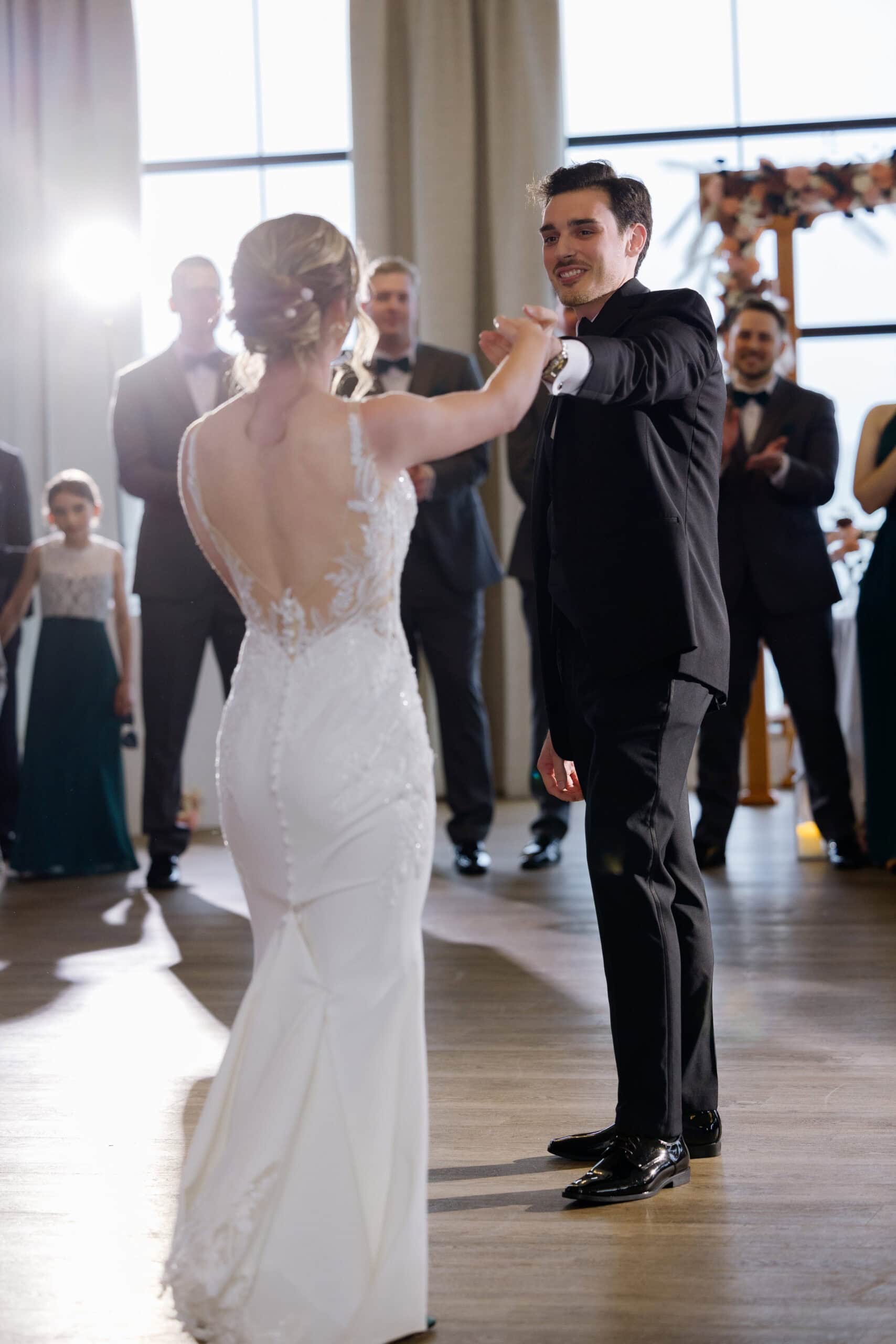Groom in black tuxedo holding hands with bride in lace-back wedding gown during first dance at Tyde at Walnut Beach venue.