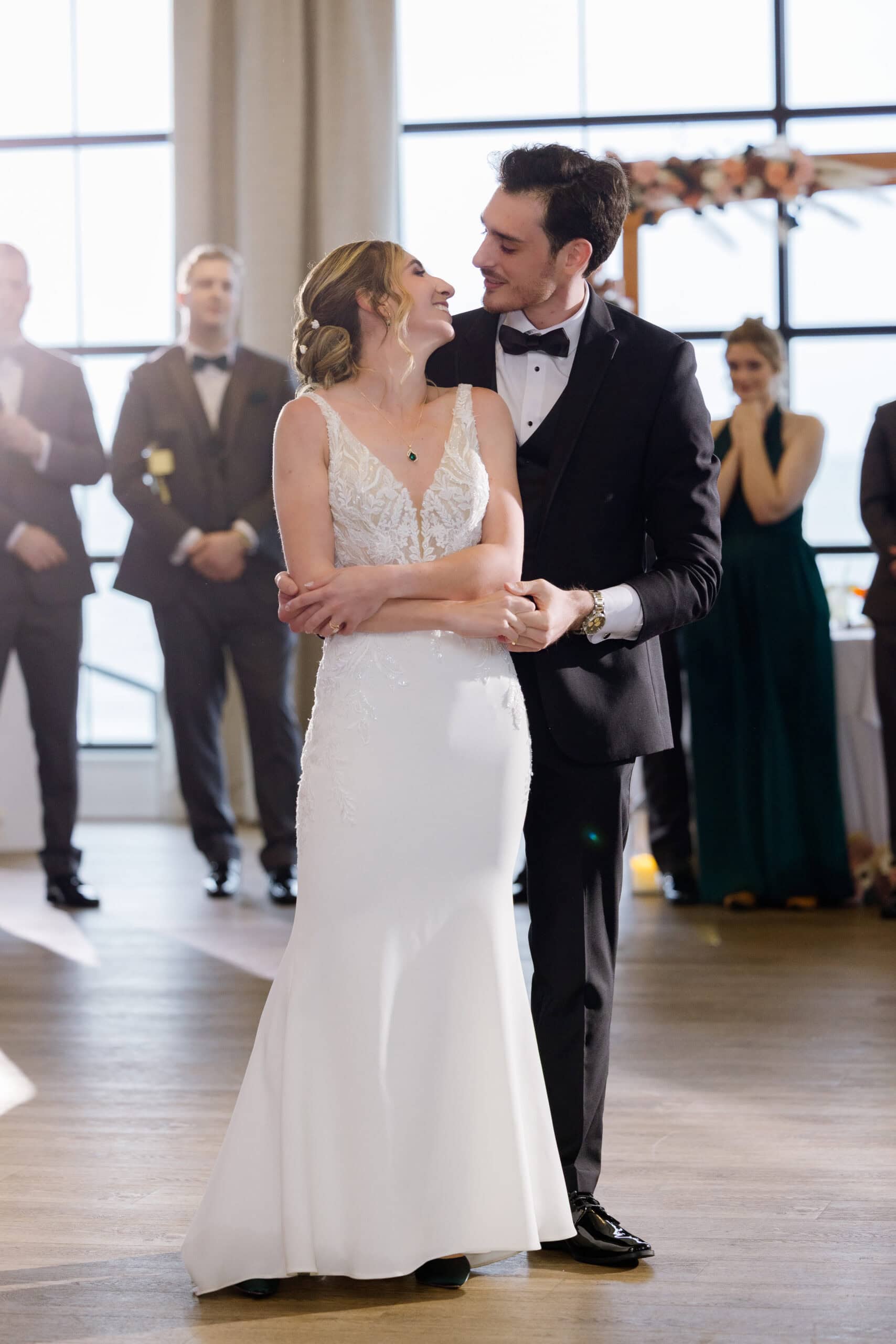 Bride in lace wedding gown and groom in black tuxedo embrace lovingly during ceremony with wedding party blurred in background at Tyde at Walnut Beach, Milford, CT.