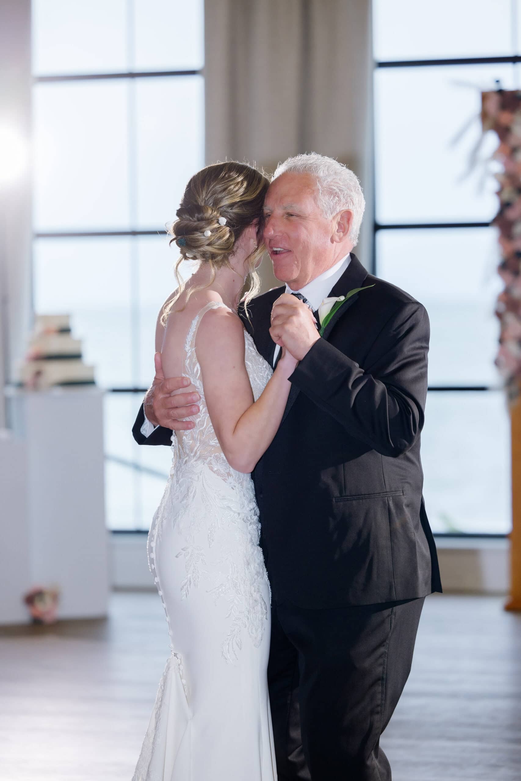 Bride in lace wedding dress dancing closely with her father in a softly lit room with large windows at Tyde at Walnut Beach, Milford, CT.