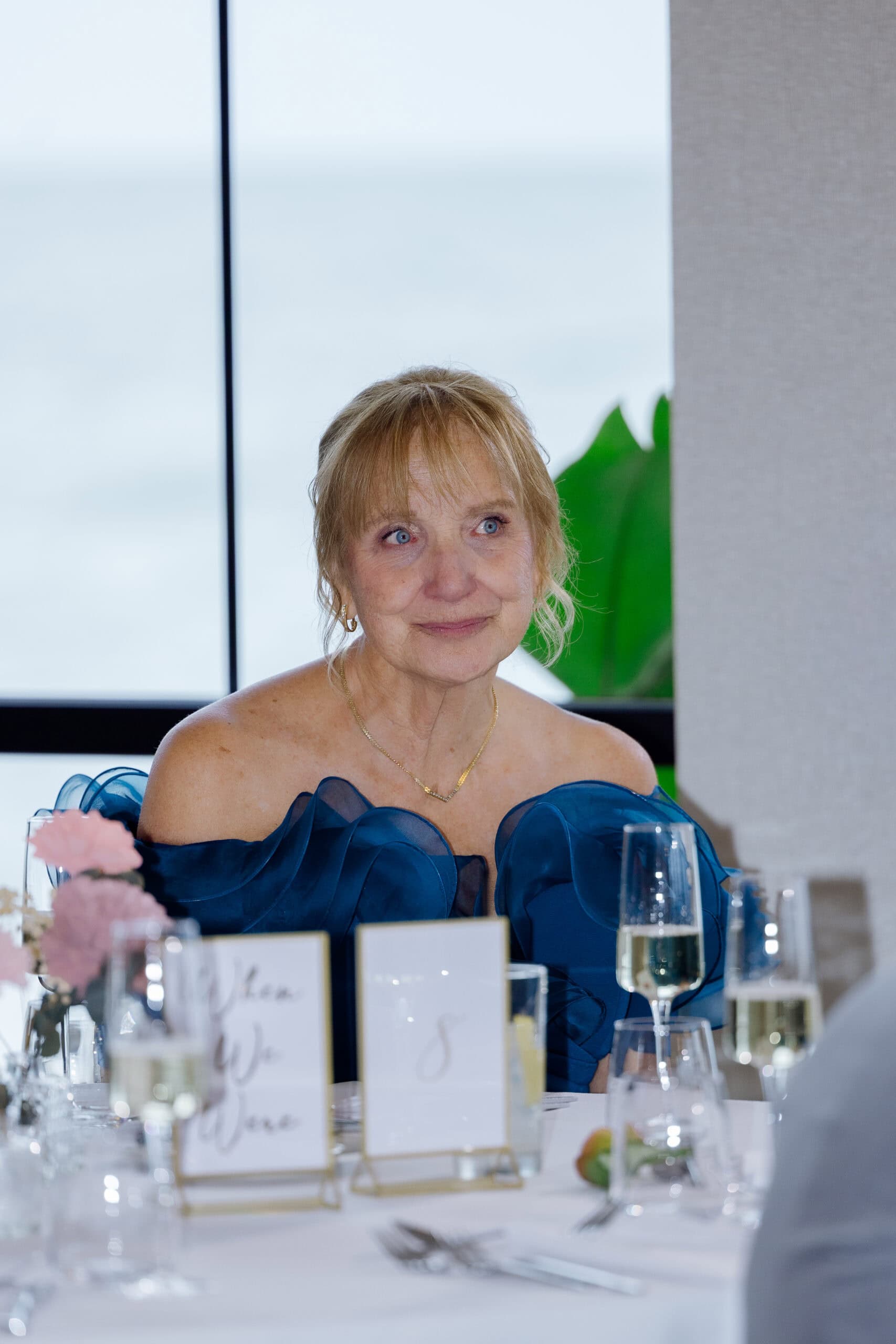 Woman in blue organza gown seated at a wedding reception table with champagne flutes and floral arrangements, Tyde at Walnut Beach, Milford, CT.