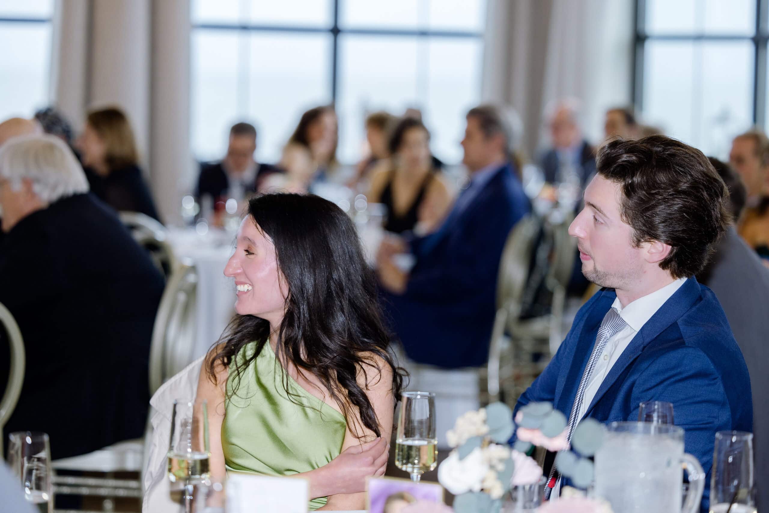 Man and woman seated at a wedding reception table, attentively listening, with blurred guests and large windows in the background at Tyde at Walnut Beach, Milford, CT.