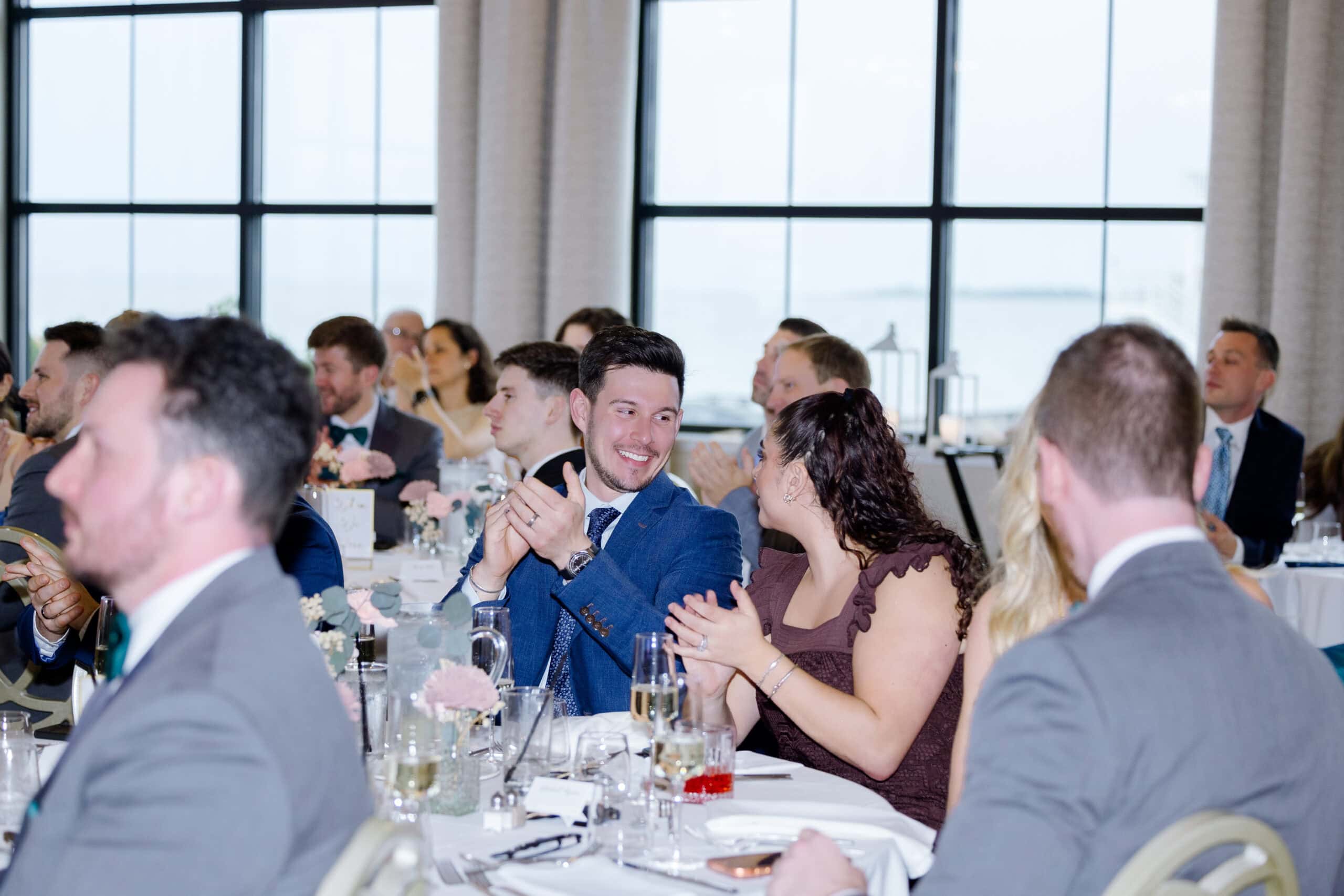 Wedding guests seated at tables clapping and conversing inside a bright venue with large windows showing a waterfront view at Tyde at Walnut Beach, Milford, CT.