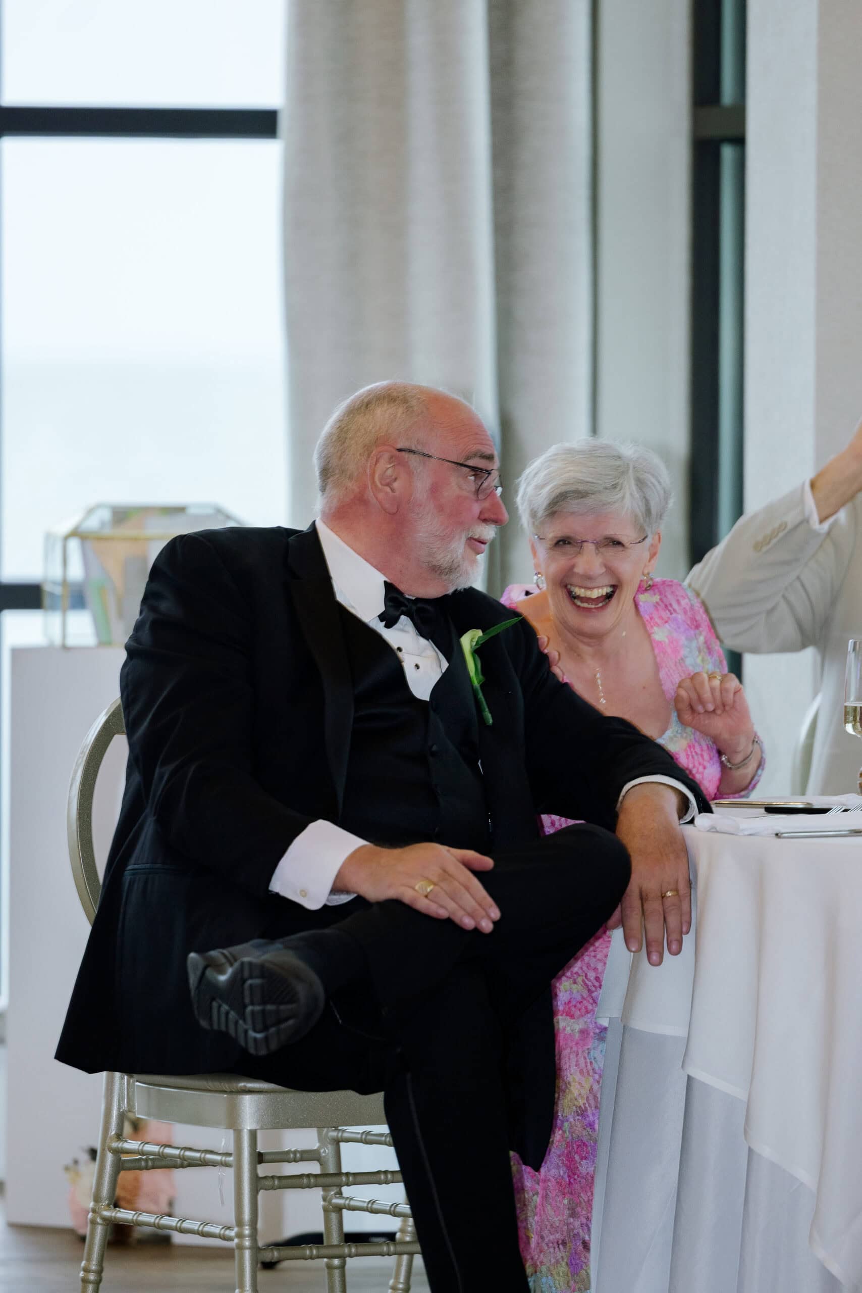 Older man in black tuxedo and woman in pink floral dress laughing together at a wedding reception table at Tyde at Walnut Beach, Milford, CT.
