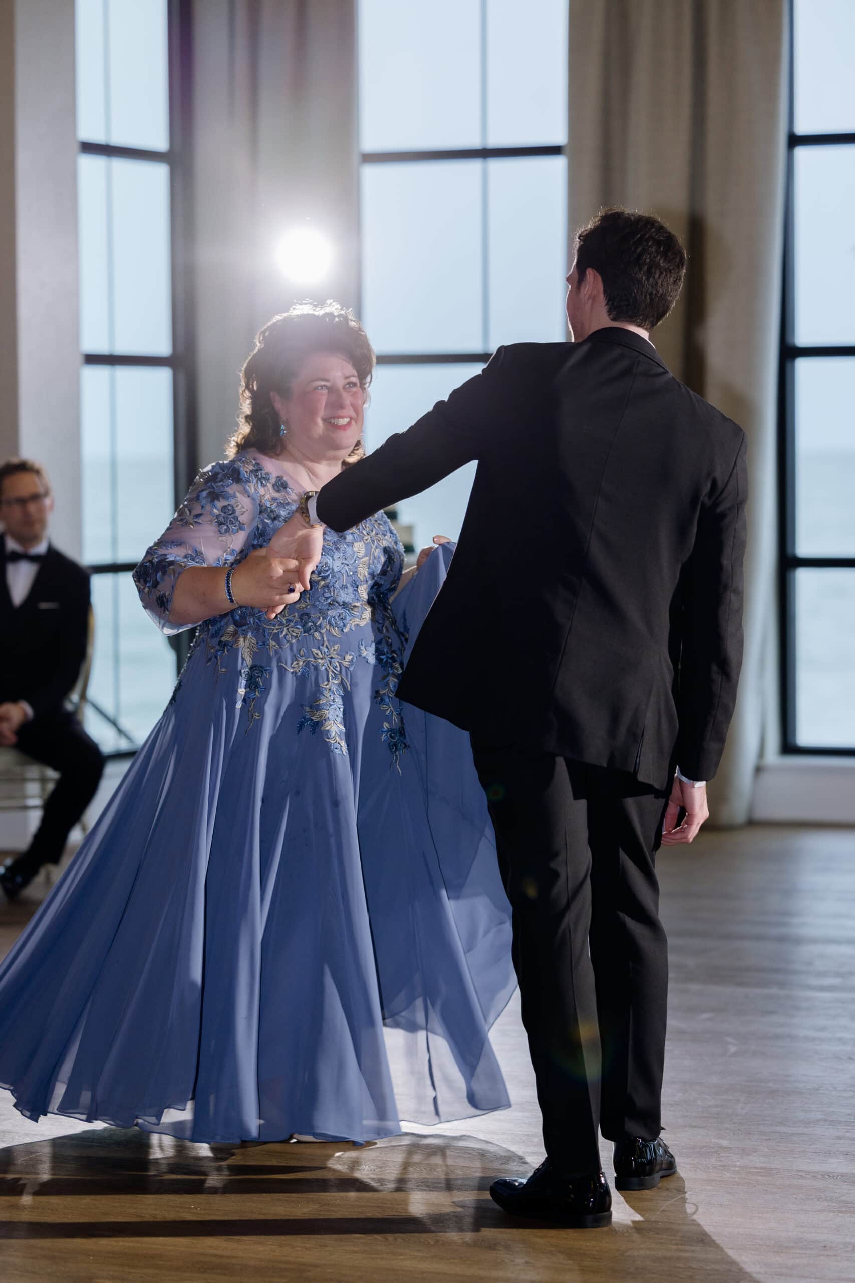 Mother in embroidered lavender gown dancing with son in black tuxedo inside a venue with large windows overlooking water at Tyde at Walnut Beach, Milford, CT.