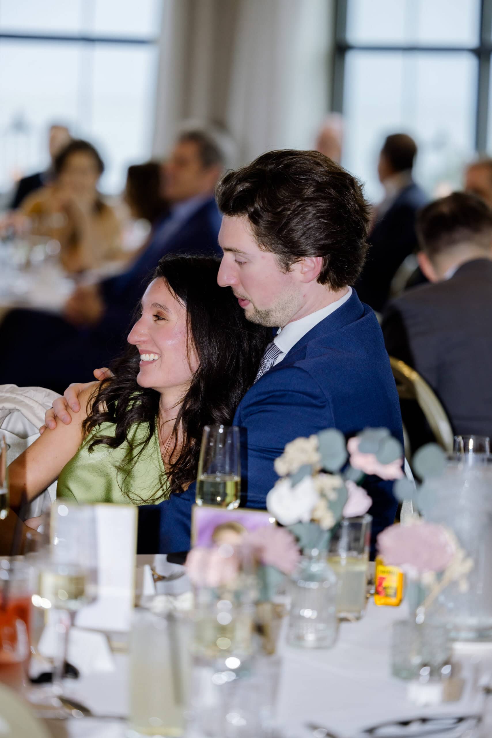 Man in navy suit gently embracing woman in green dress at wedding reception with blurred guests and window background.