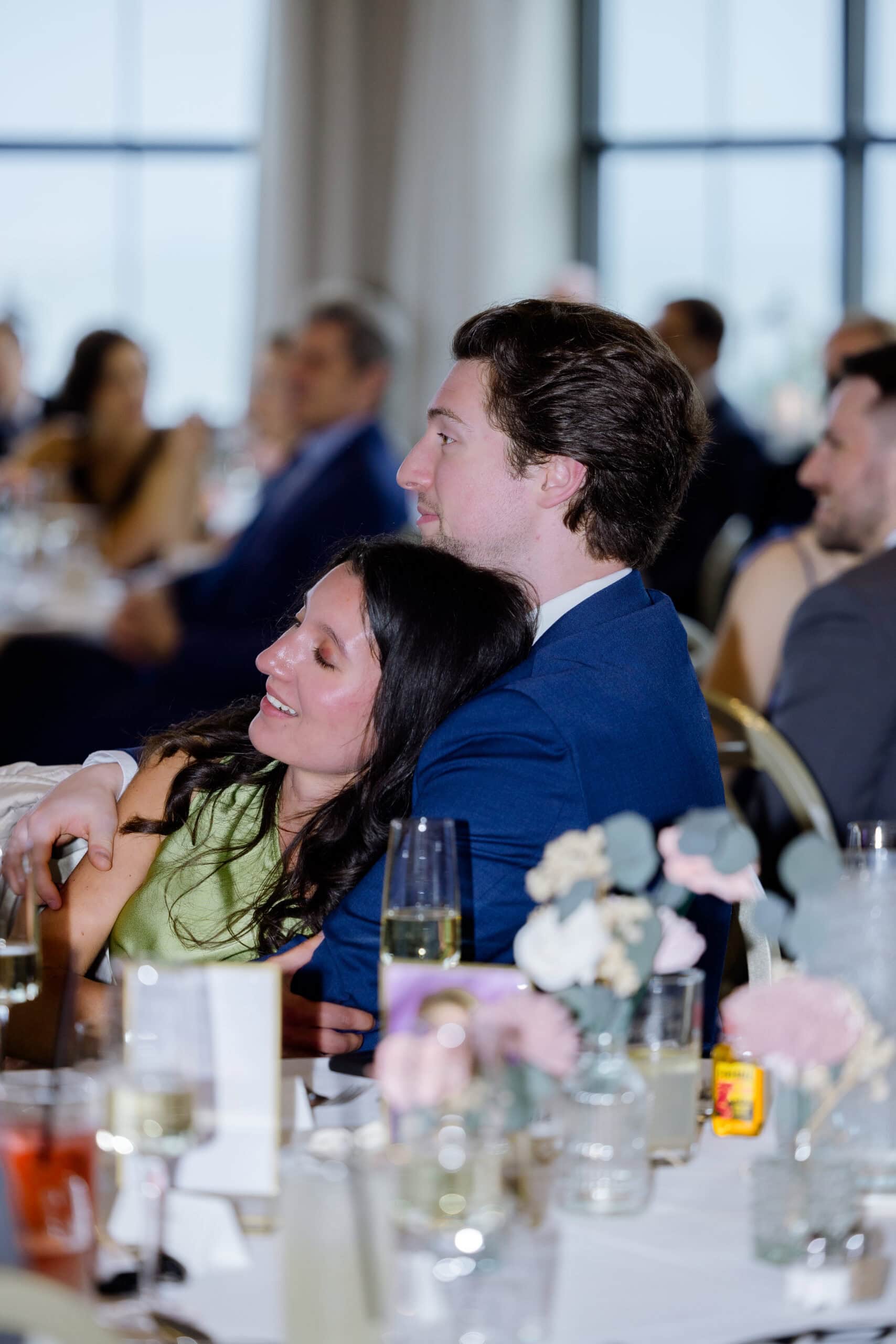 Couple embracing at wedding reception with blurred guests and floral table decor in background at Tyde at Walnut Beach, Milford, CT.