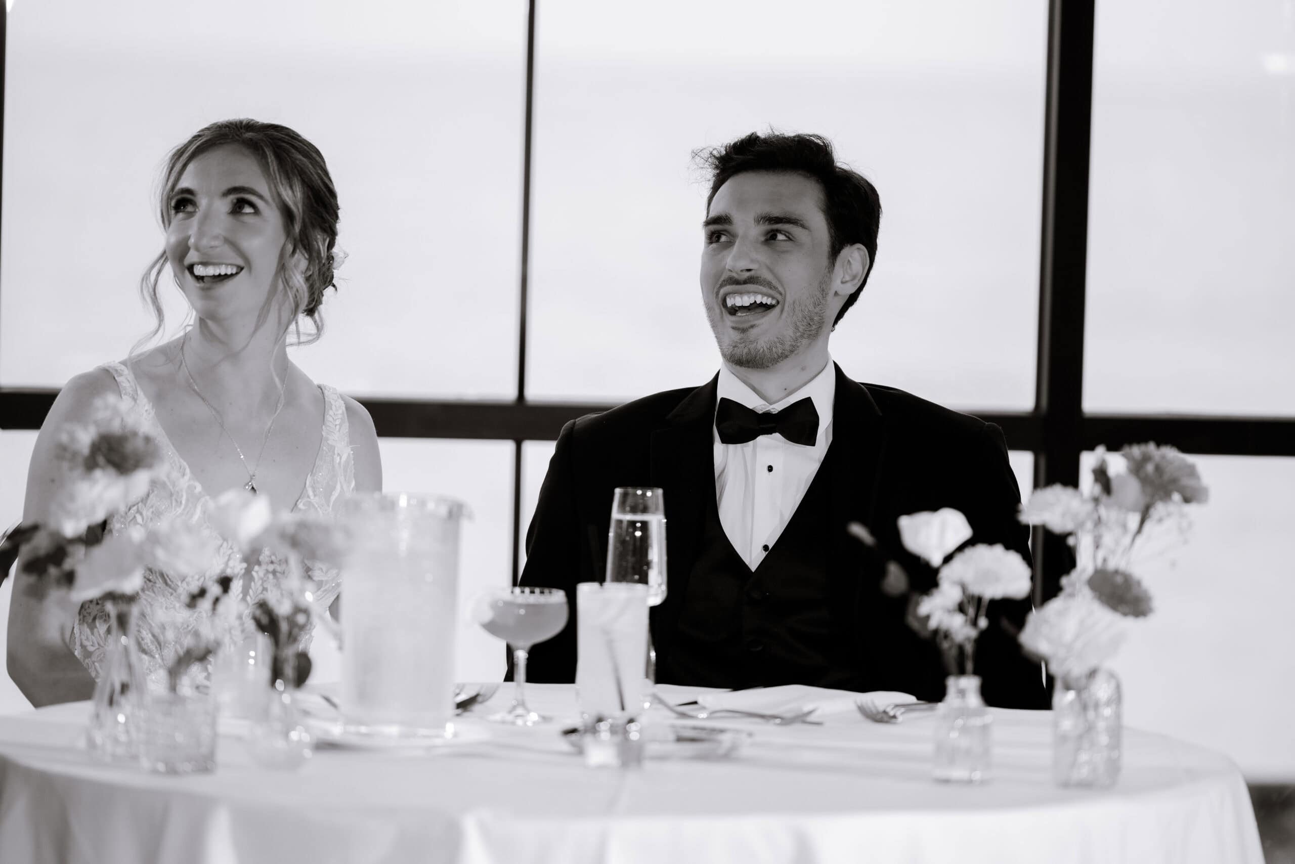 Black and white photo of a bride and groom laughing together seated at a table with floral centerpieces and glassware, with large windows in the background at Tyde at Walnut Beach, Milford, CT.