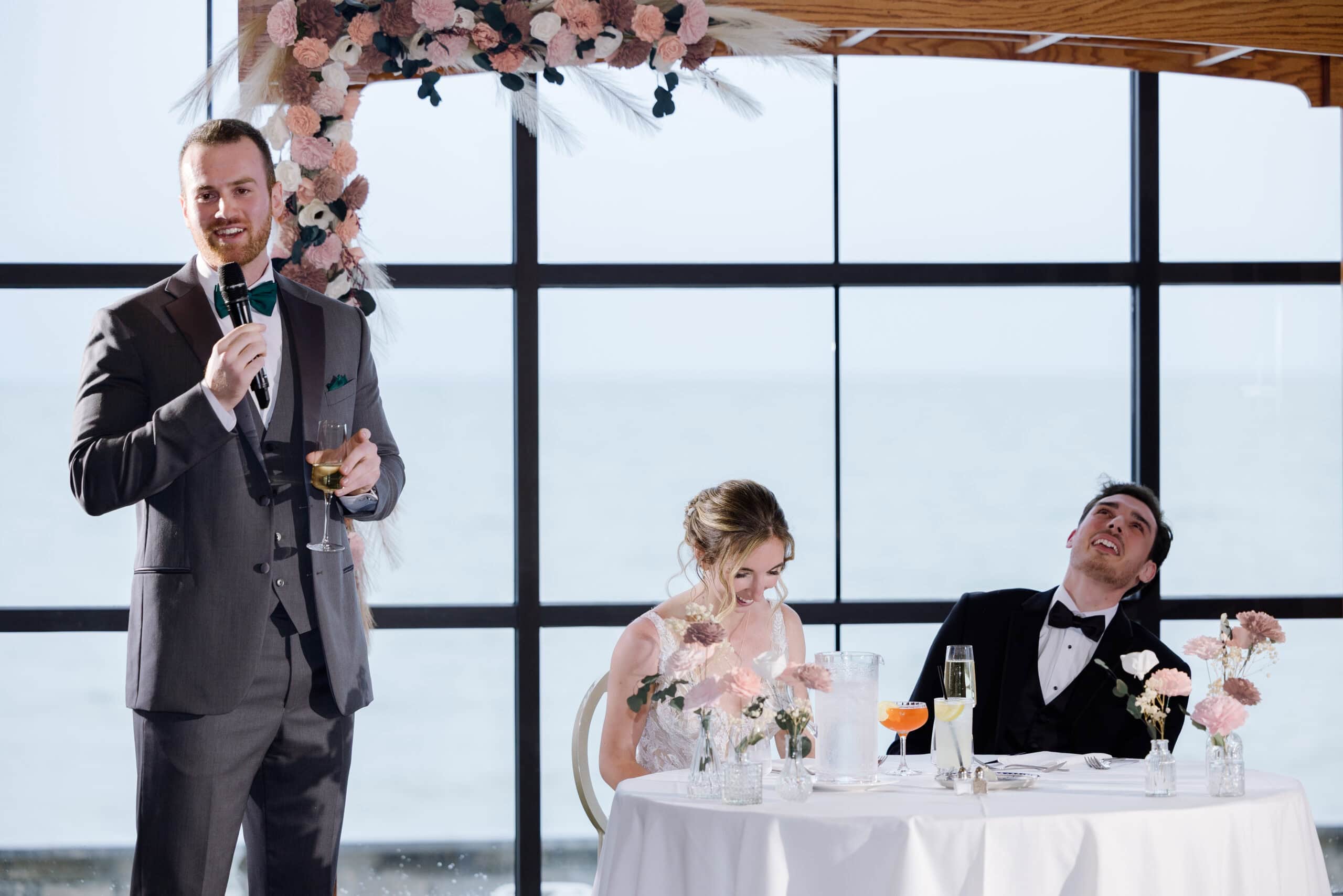 Man in gray suit giving a wedding toast holding a microphone and champagne glass, bride and groom seated at a table with pink floral arrangements and ocean visible through large windows behind them.