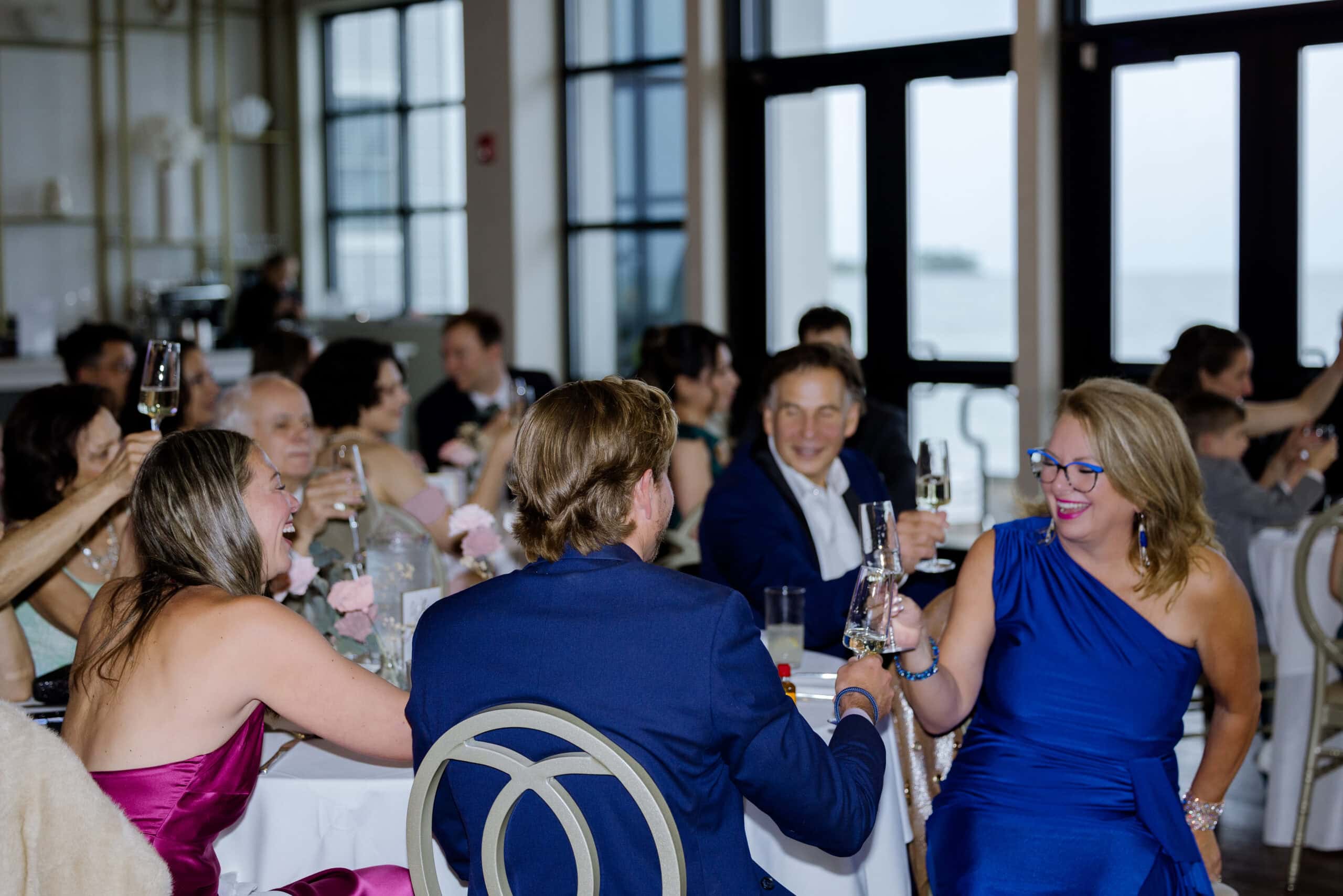Guests seated around a table raising champagne glasses in toast inside a bright venue with large windows overlooking water.