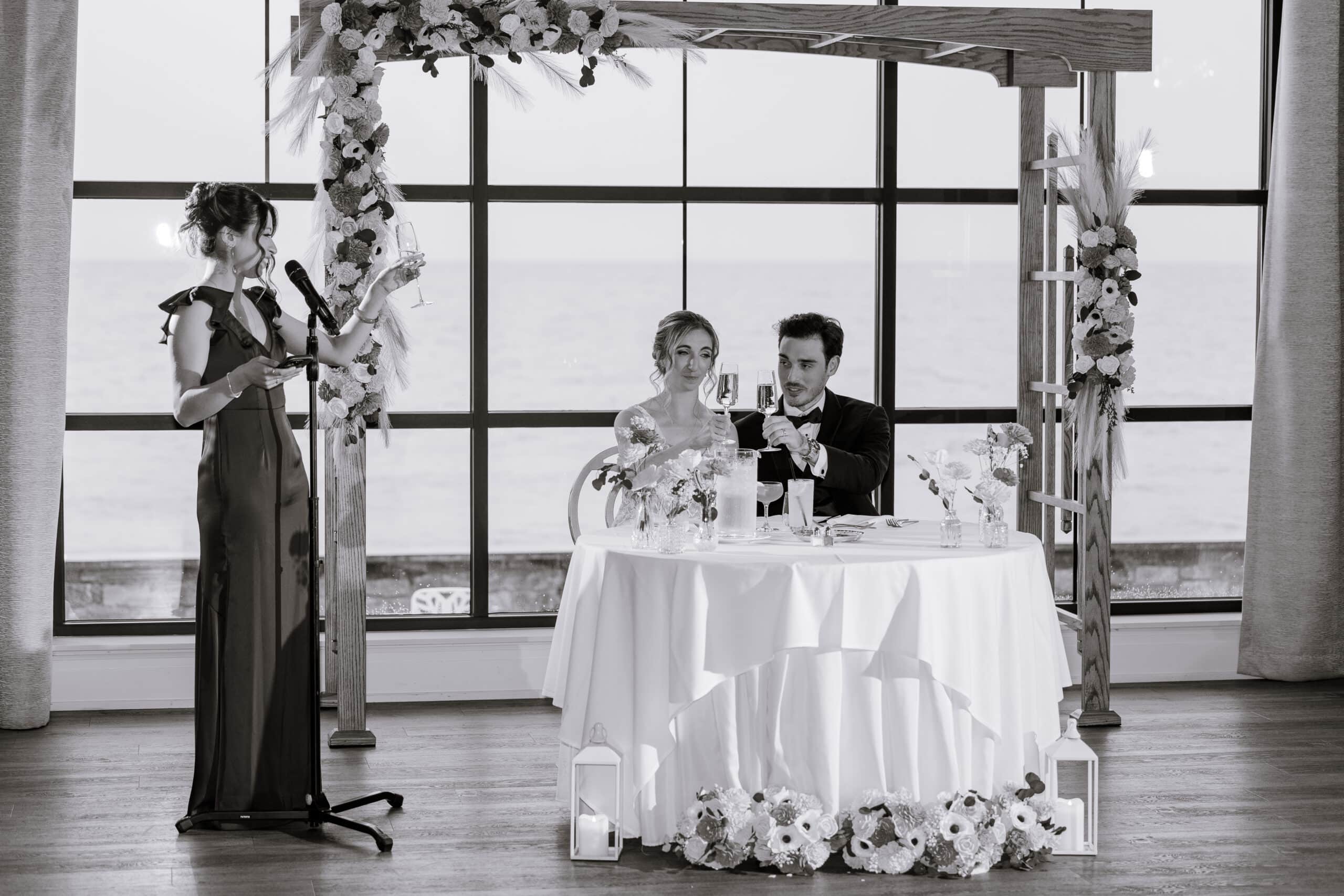 Woman giving a wedding toast with a microphone, bride and groom seated at a table adorned with flowers, ocean visible through large windows.