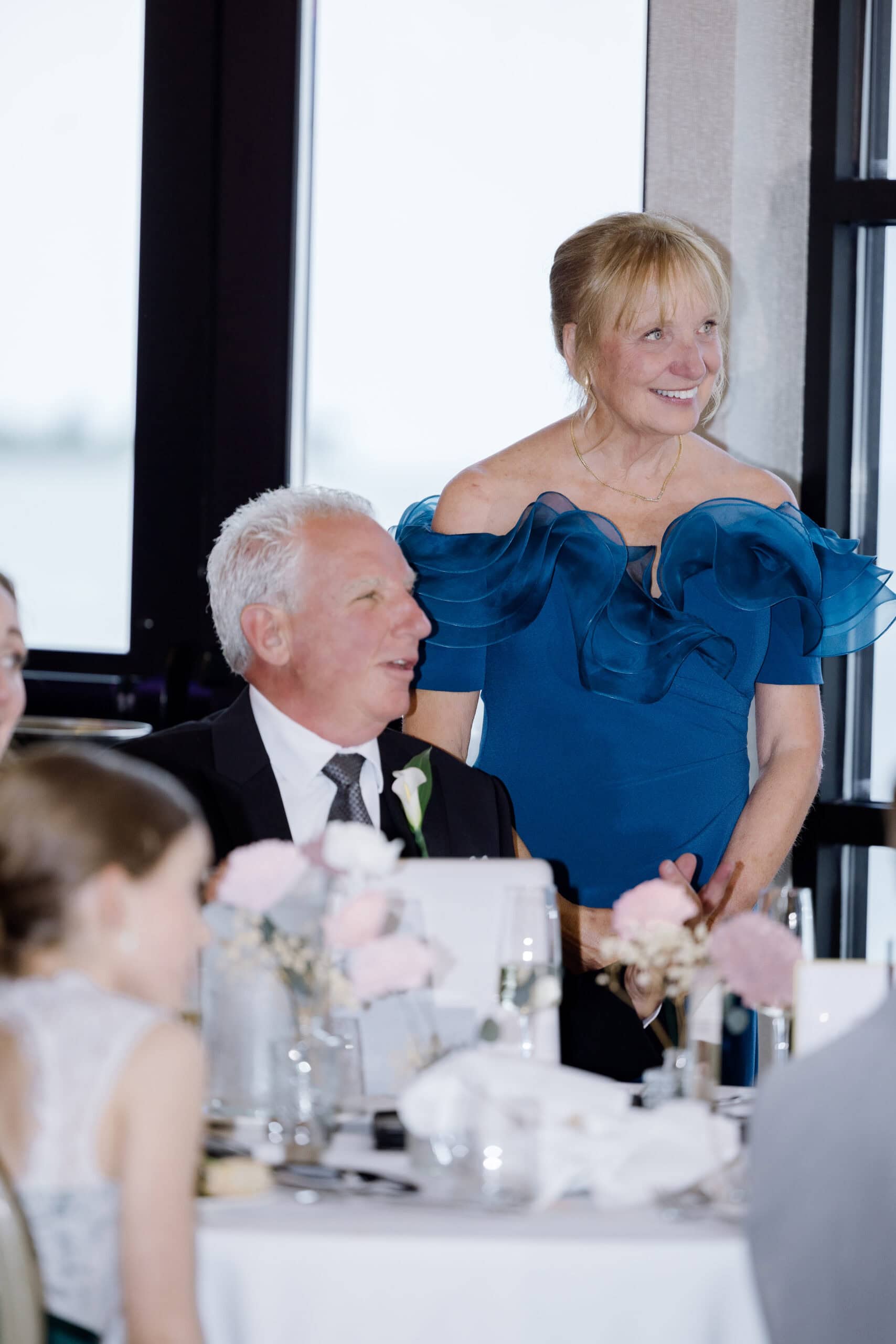 Woman in an elegant blue off-shoulder dress standing and smiling beside a seated man in a black suit at a wedding reception table with floral arrangements.