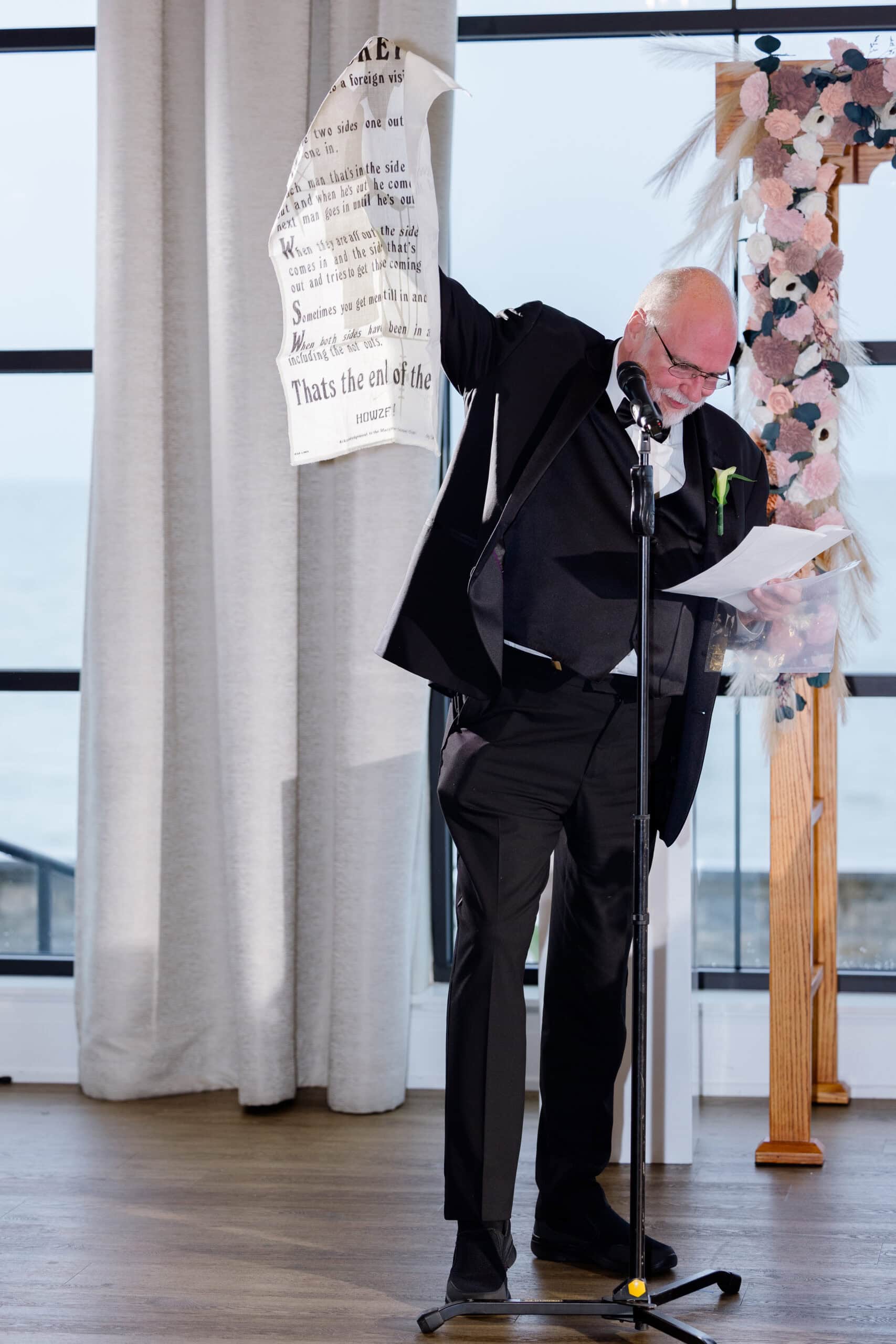 Man in black suit and glasses speaking into microphone, holding a printed linen cloth, standing near floral arch indoors with ocean view