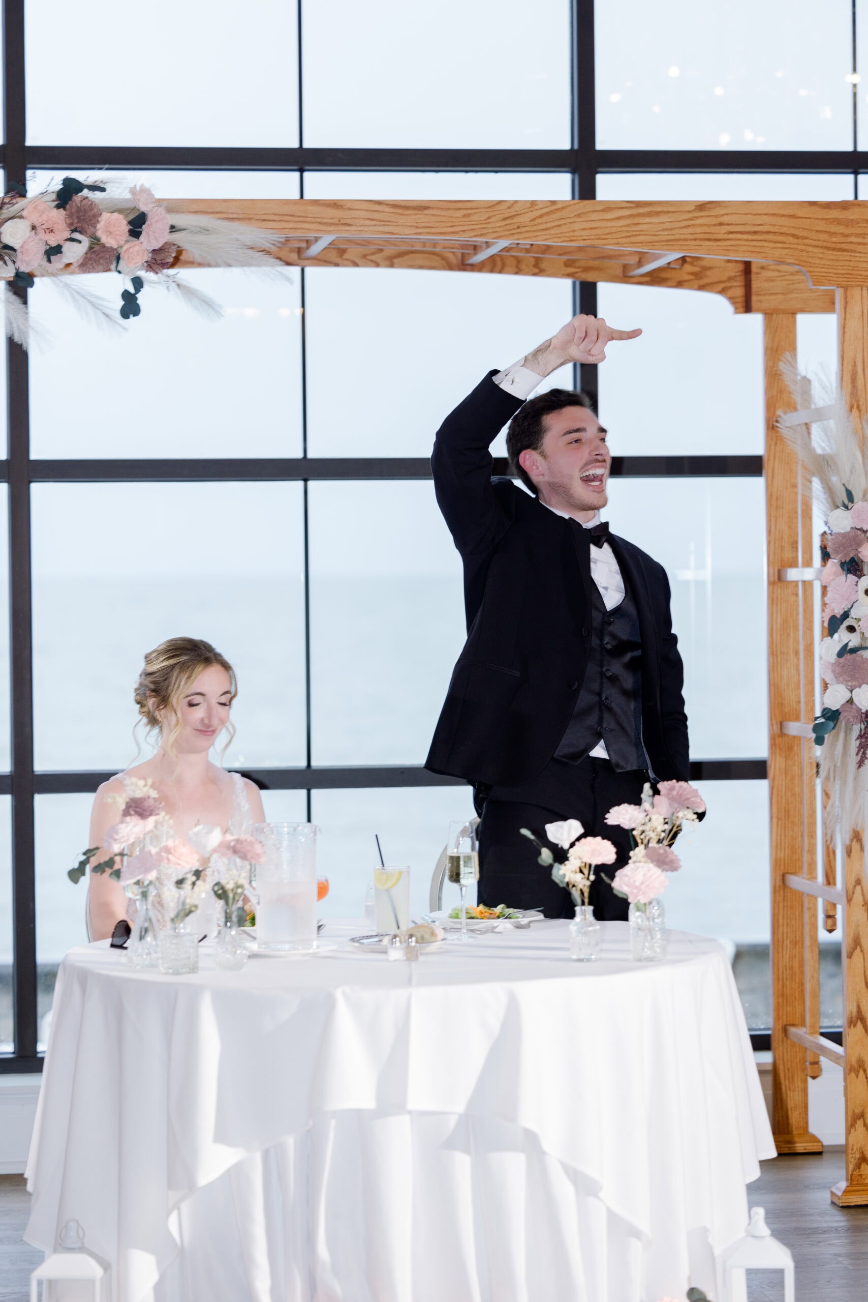 Groom in black tuxedo raising hand and speaking during toast at wedding reception with bride seated beside him, floral arch and large windows overlooking water in background at Tyde at Walnut Beach, Milford, CT.