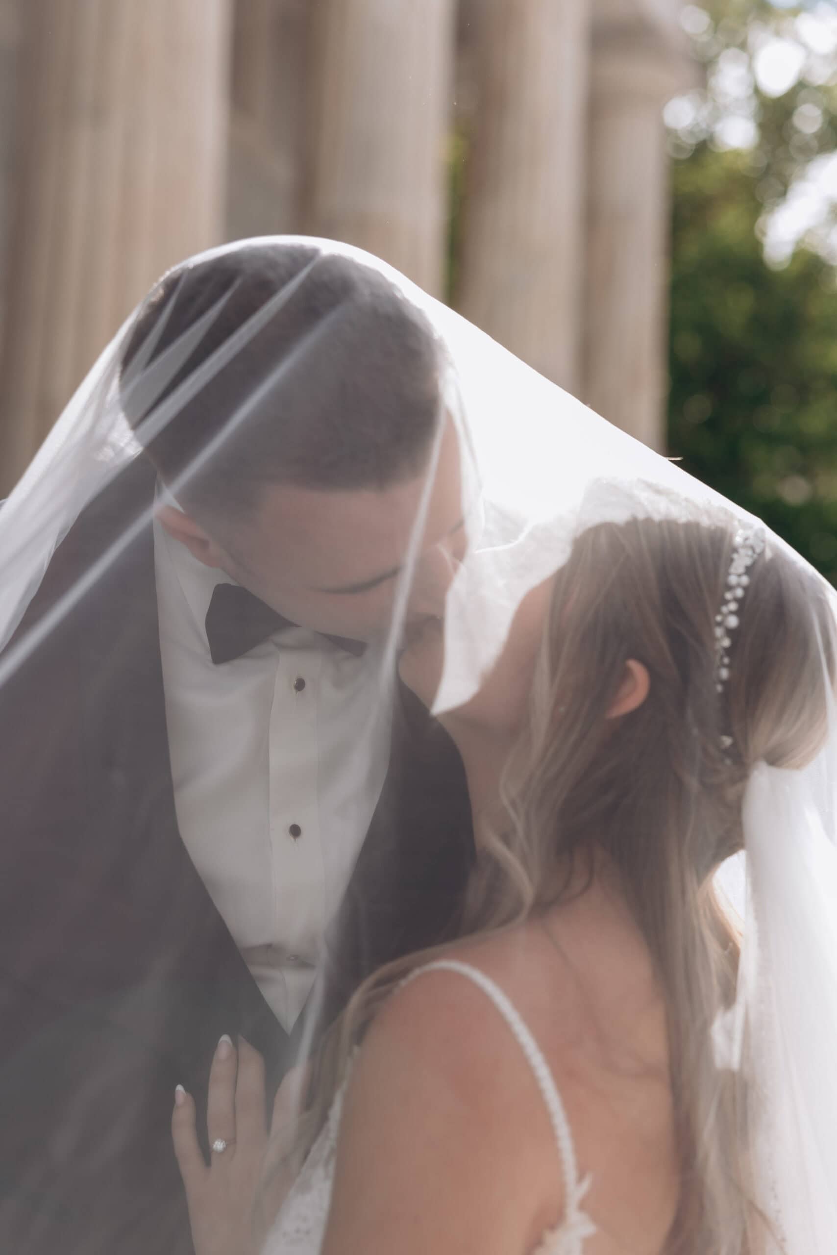 Close-up of bride and groom kissing under a sheer veil with classical columns and greenery blurred in the background at Union Trust, Philadelphia.