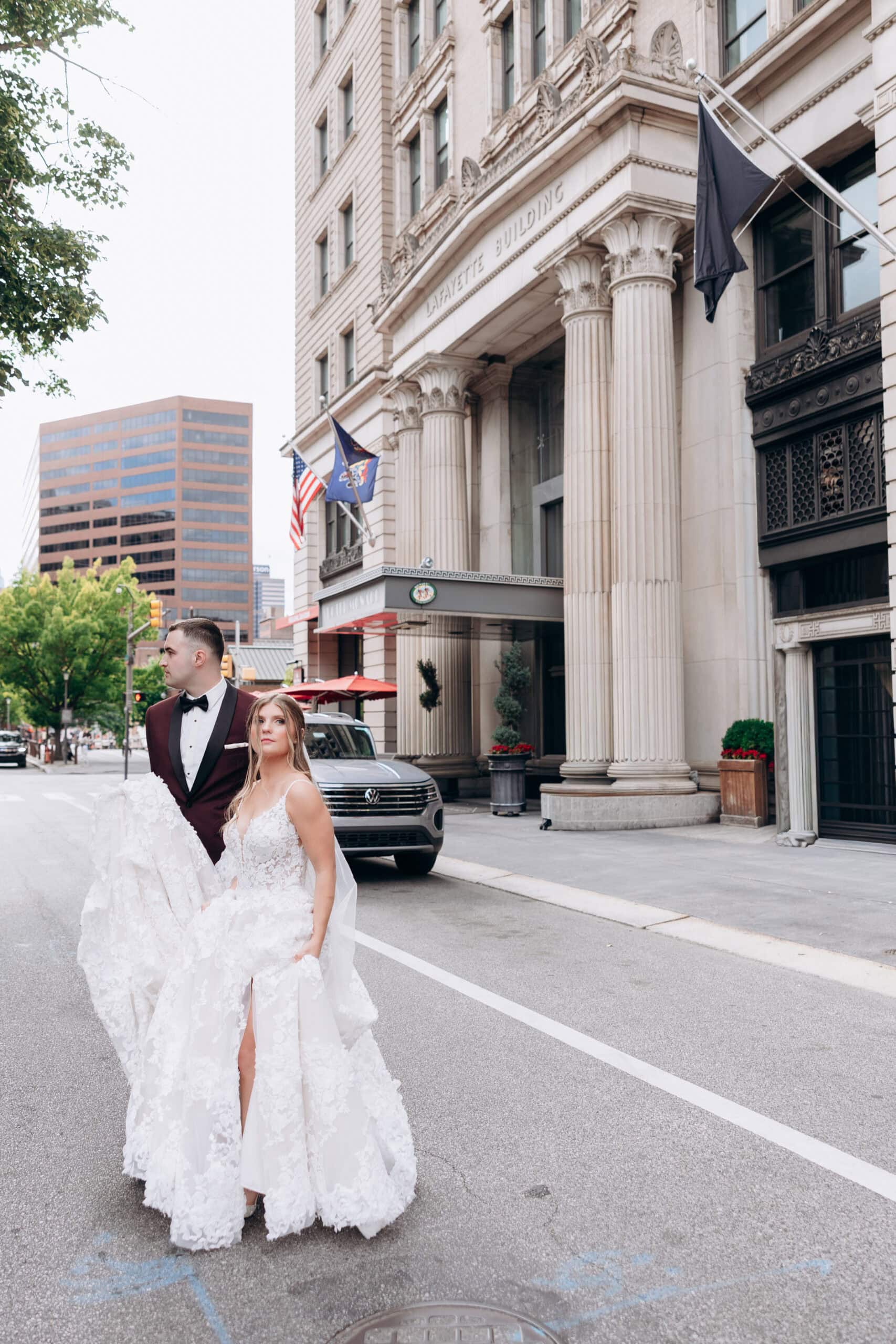 Bride in floral lace gown and groom in burgundy tuxedo standing on street near Lafayette Building with classical columns in Philadelphia.