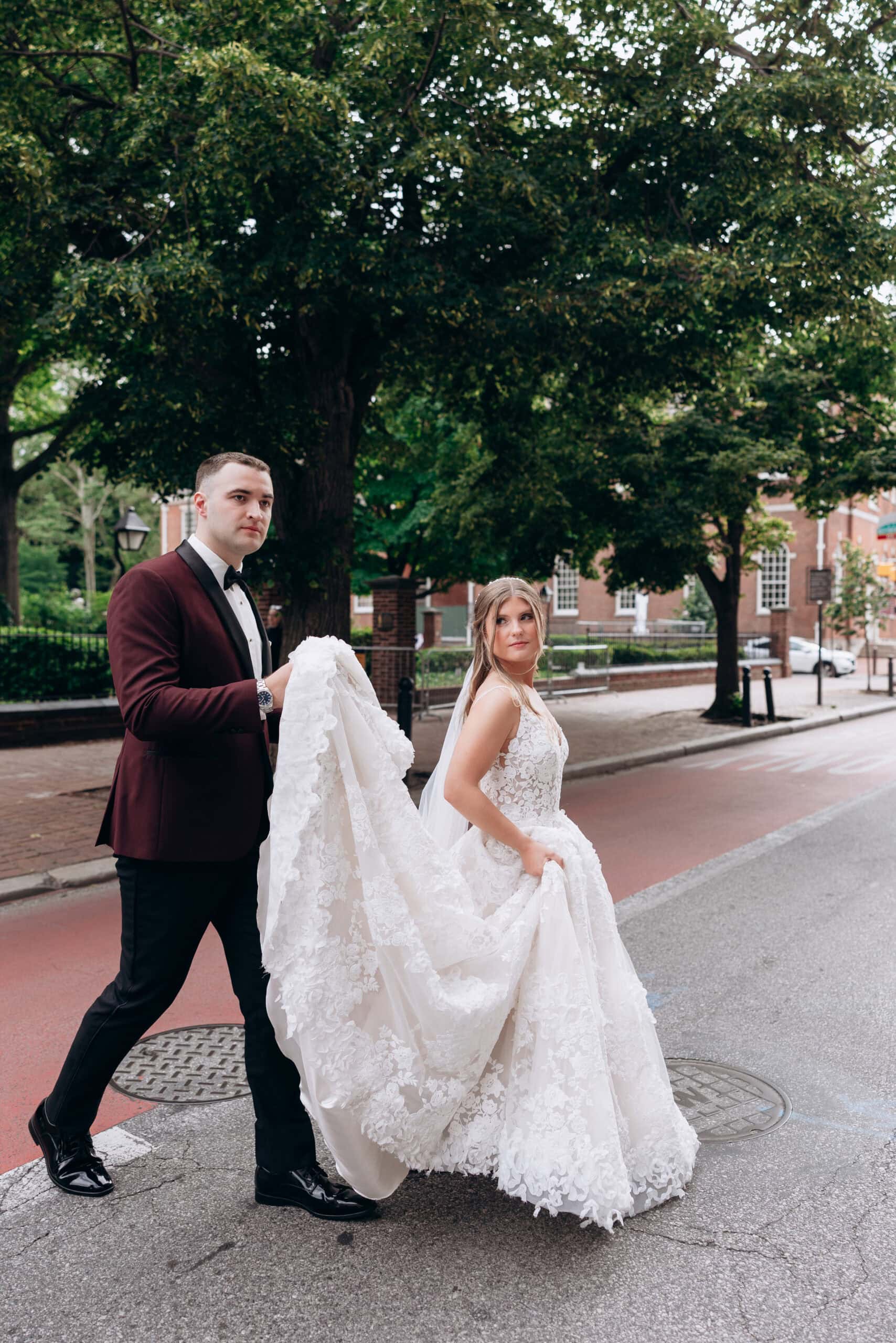 Groom in burgundy tuxedo holding the lace train of bride’s detailed white dress on a city street with trees in the background at Union Trust, Philadelphia.