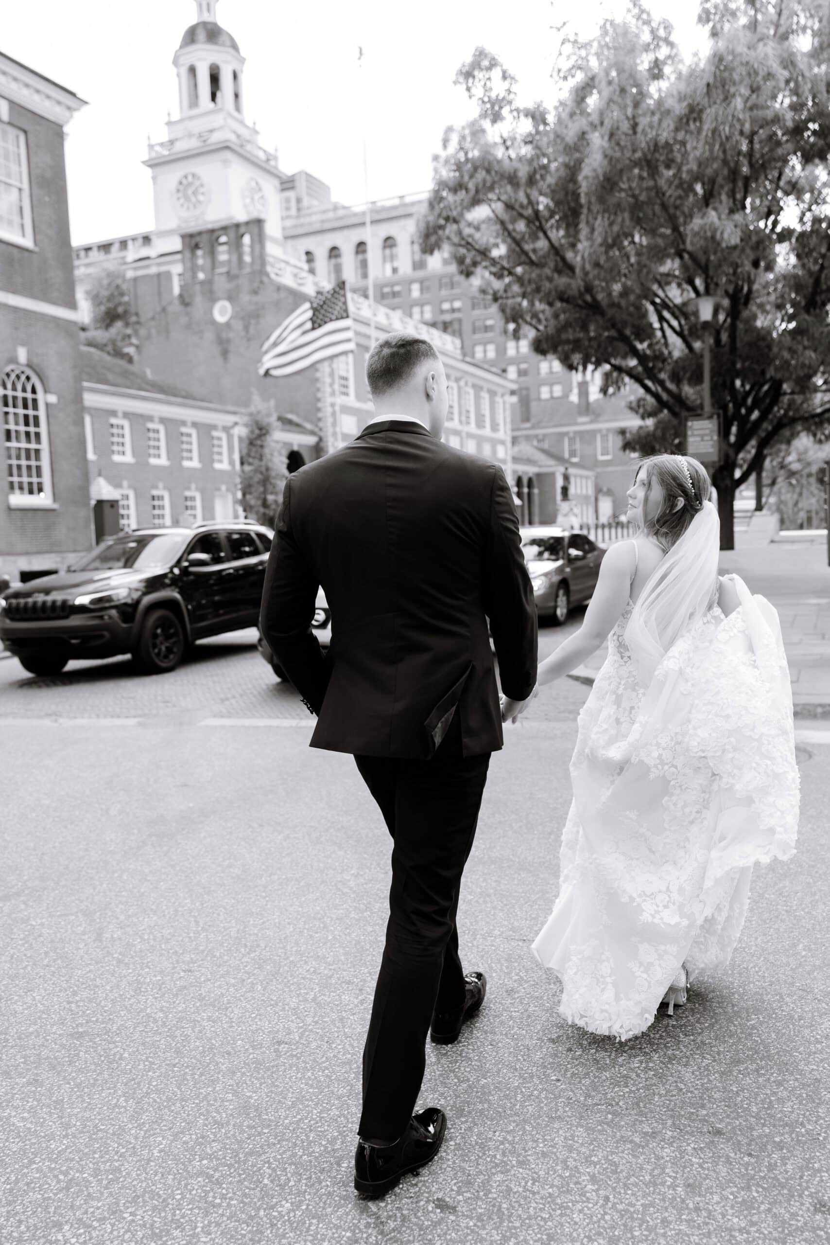 Black-and-white photo of a bride and groom walking hand-in-hand near historic Union Trust building in Philadelphia, PA, with trees and cars in background.
