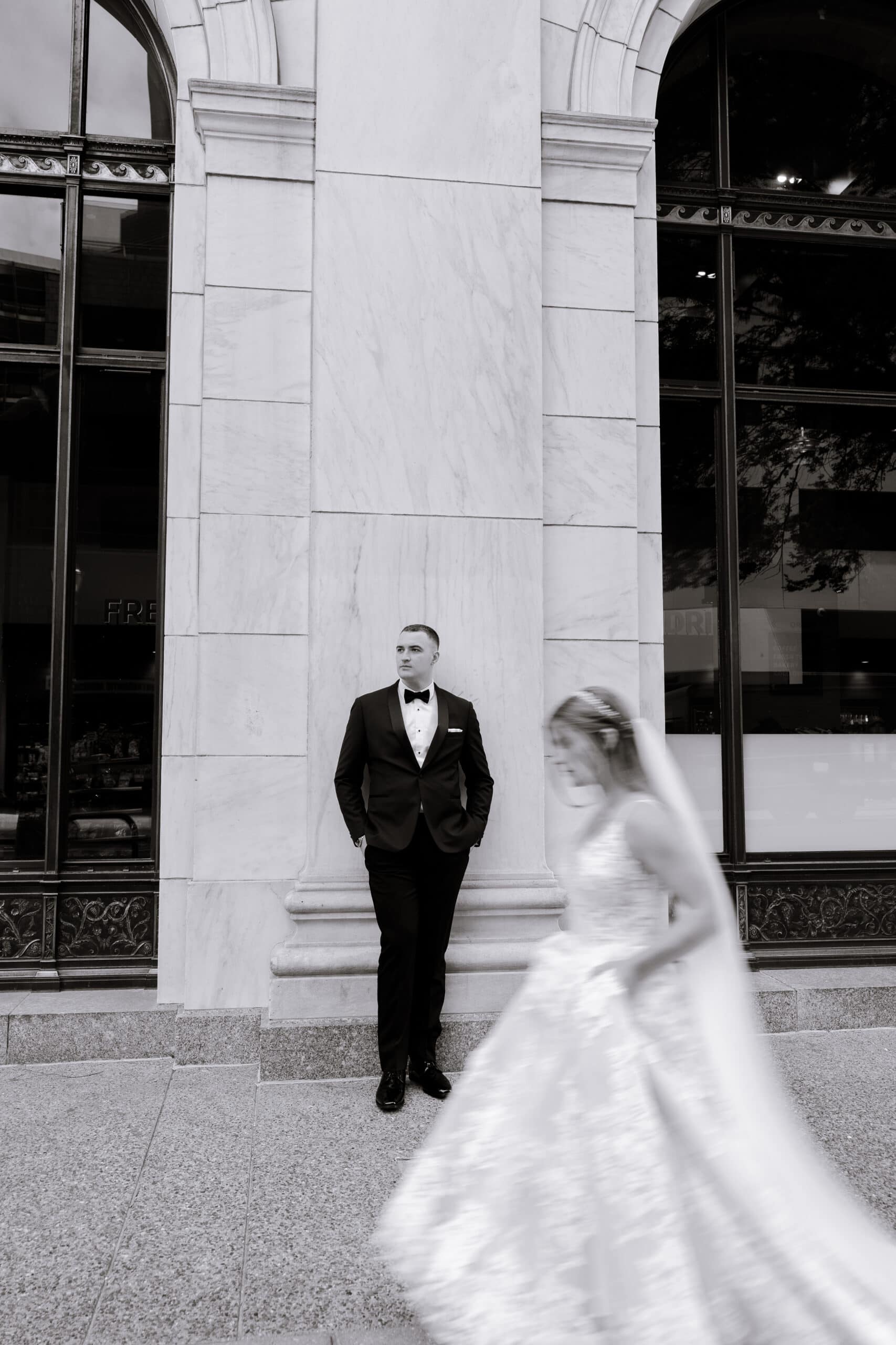 Groom in tuxedo standing against marble wall with arched windows, bride in wedding dress walking past in blurred motion at Union Trust, Philadelphia.