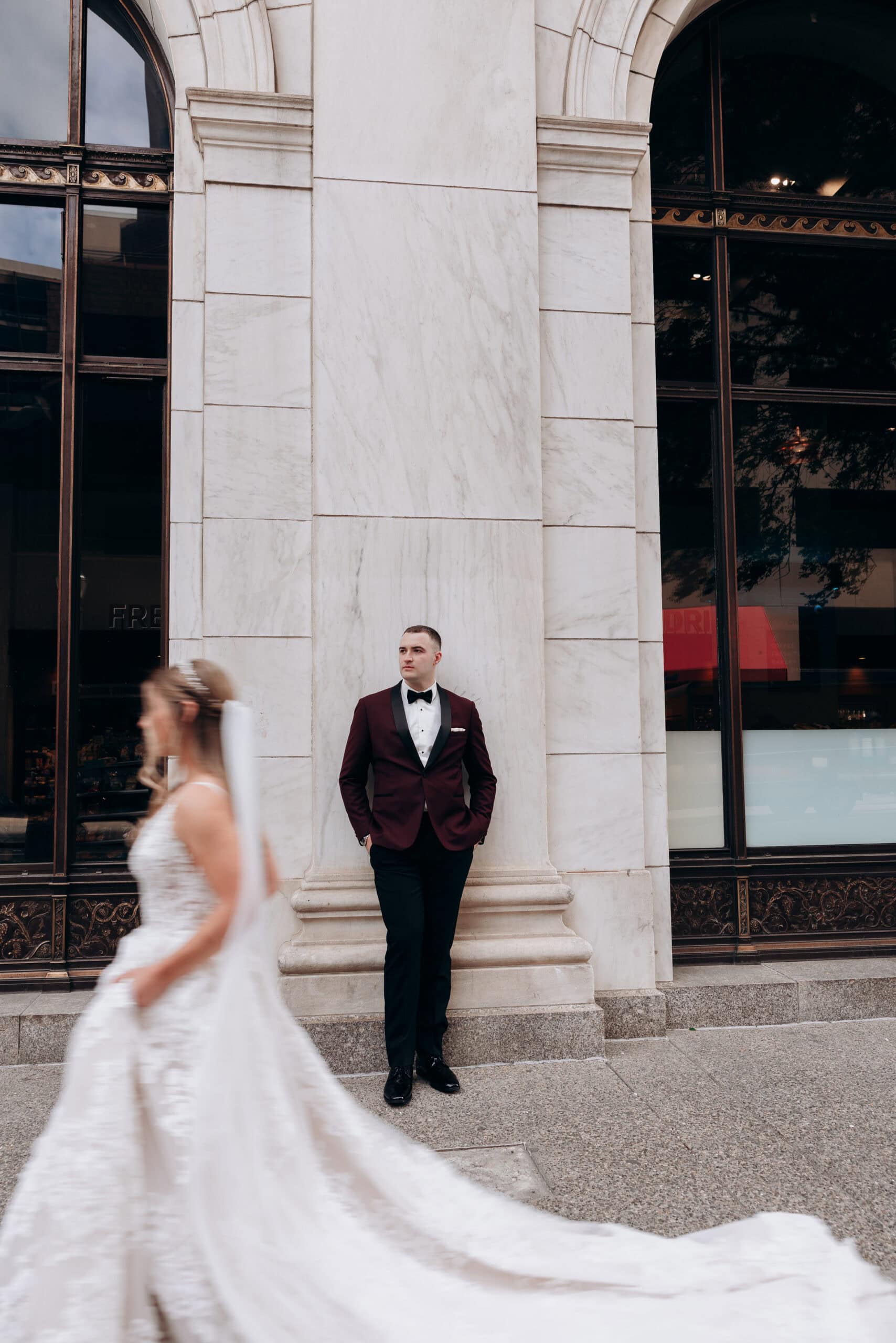 Groom in burgundy tuxedo leaning against marble wall with blurred bride walking past in white wedding gown at Union Trust, Philadelphia.