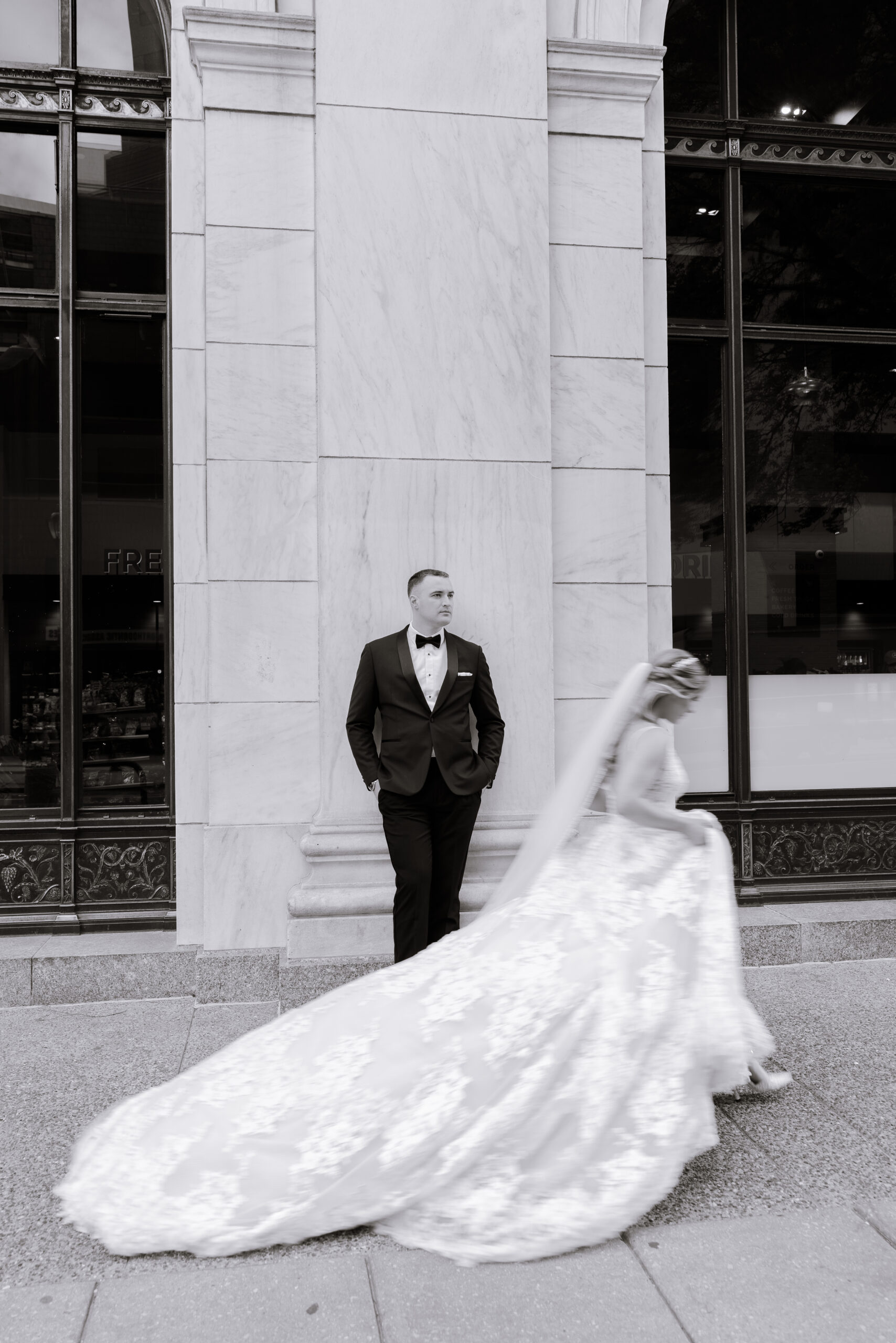 Groom in black tuxedo leaning against marble wall, bride in lace wedding dress with long train walking past in motion blur at Union Trust, Philadelphia.
